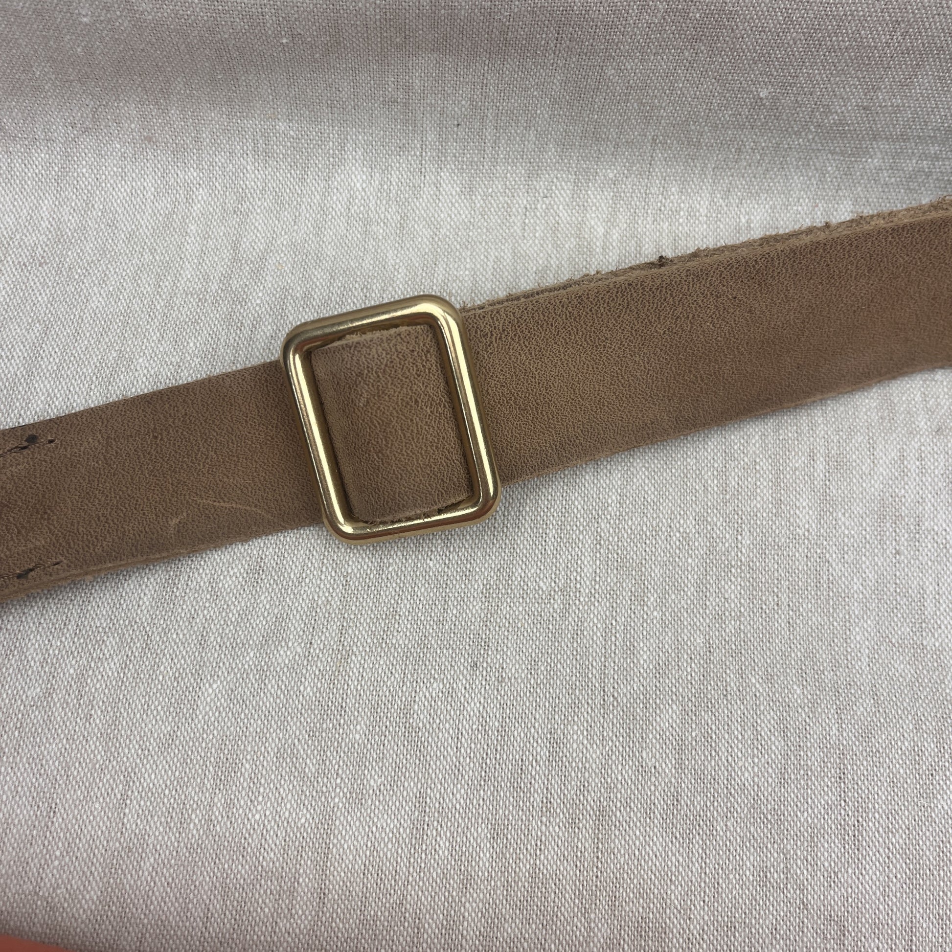 A close-up of a brown leather strap with a square gold-toned metal buckle, laid diagonally on a light gray textured fabric background.