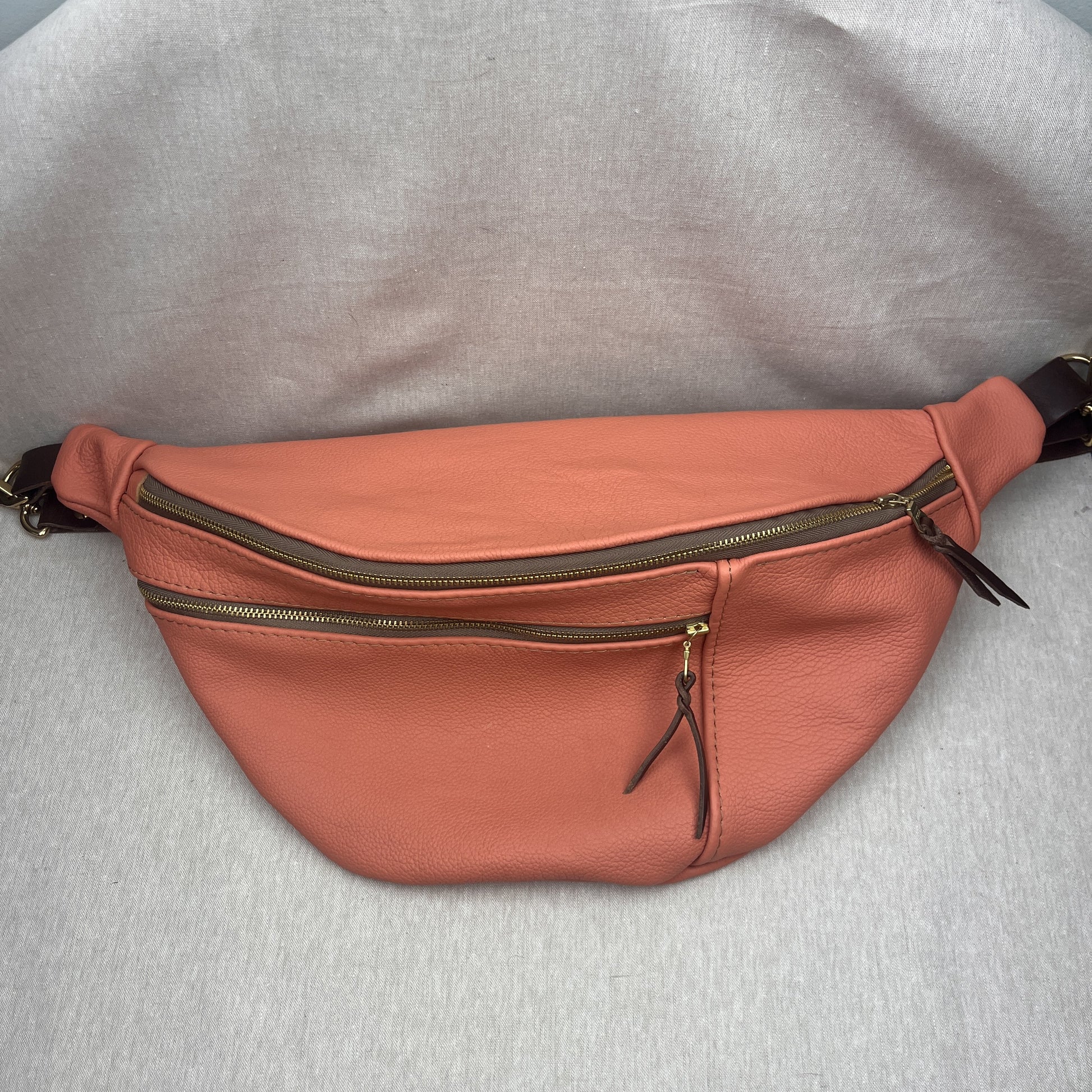 A peach-colored leather fanny pack with multiple zippered compartments and a dark strap, laid flat on a light gray textured fabric background.