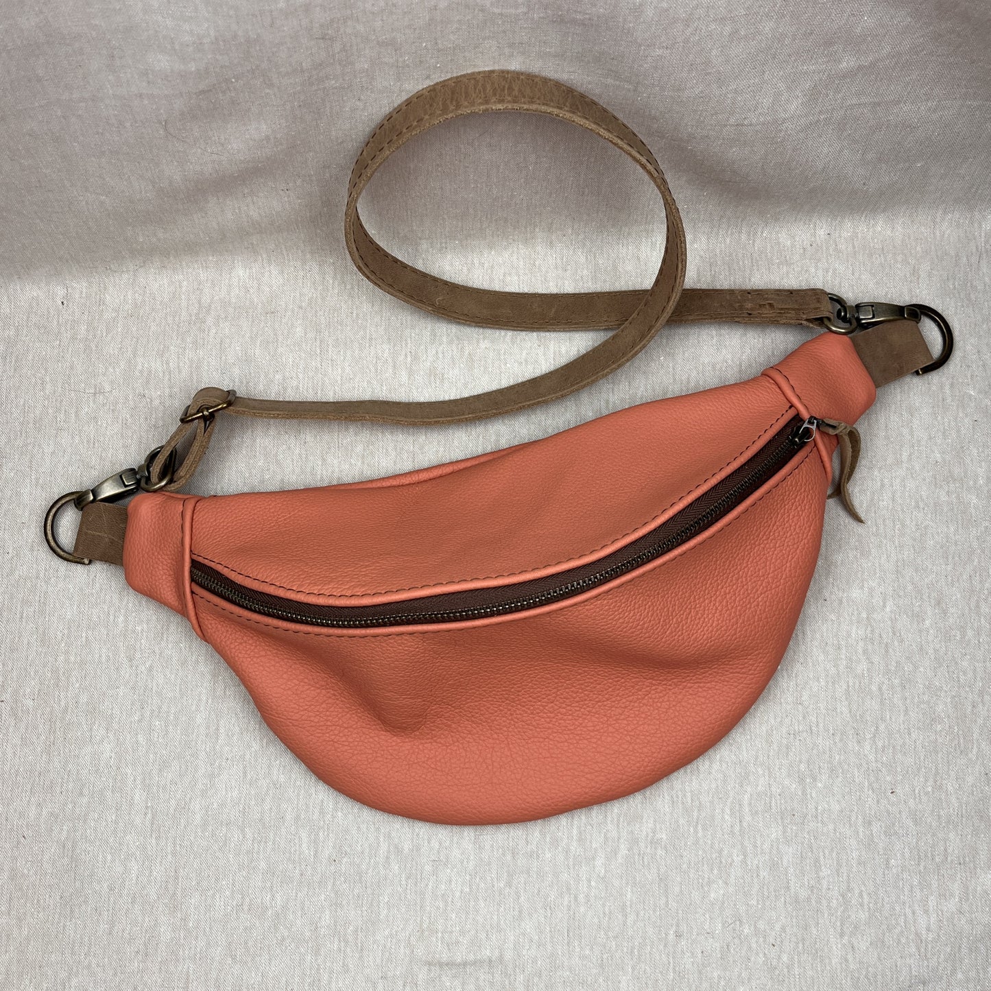 A curved fanny pack in peachy orange pebbled leather with a brown strap and metal hardware, laid flat on a light gray fabric background.