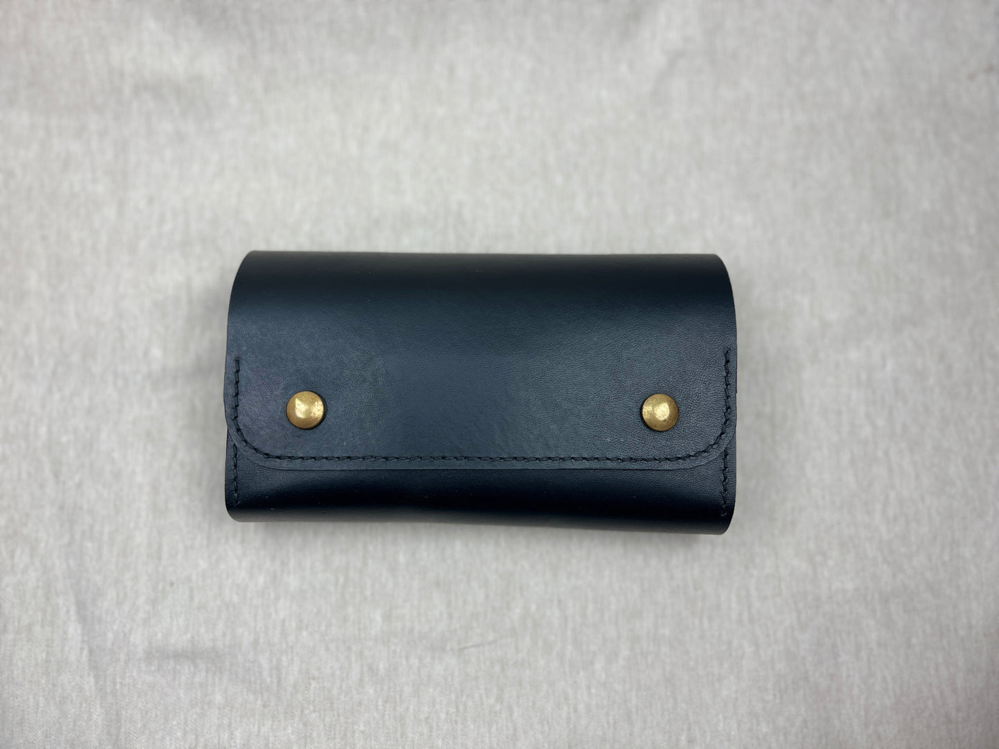 A black cigar case with two gold-tone snap buttons, shown flat on a light gray textured fabric background.