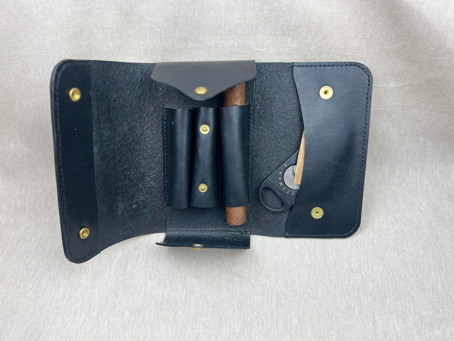 A black leather cigar accessory organizer laid open on a light gray fabric background, containing a cigar in a roll holder, a small pouch, and a cigar cutter.