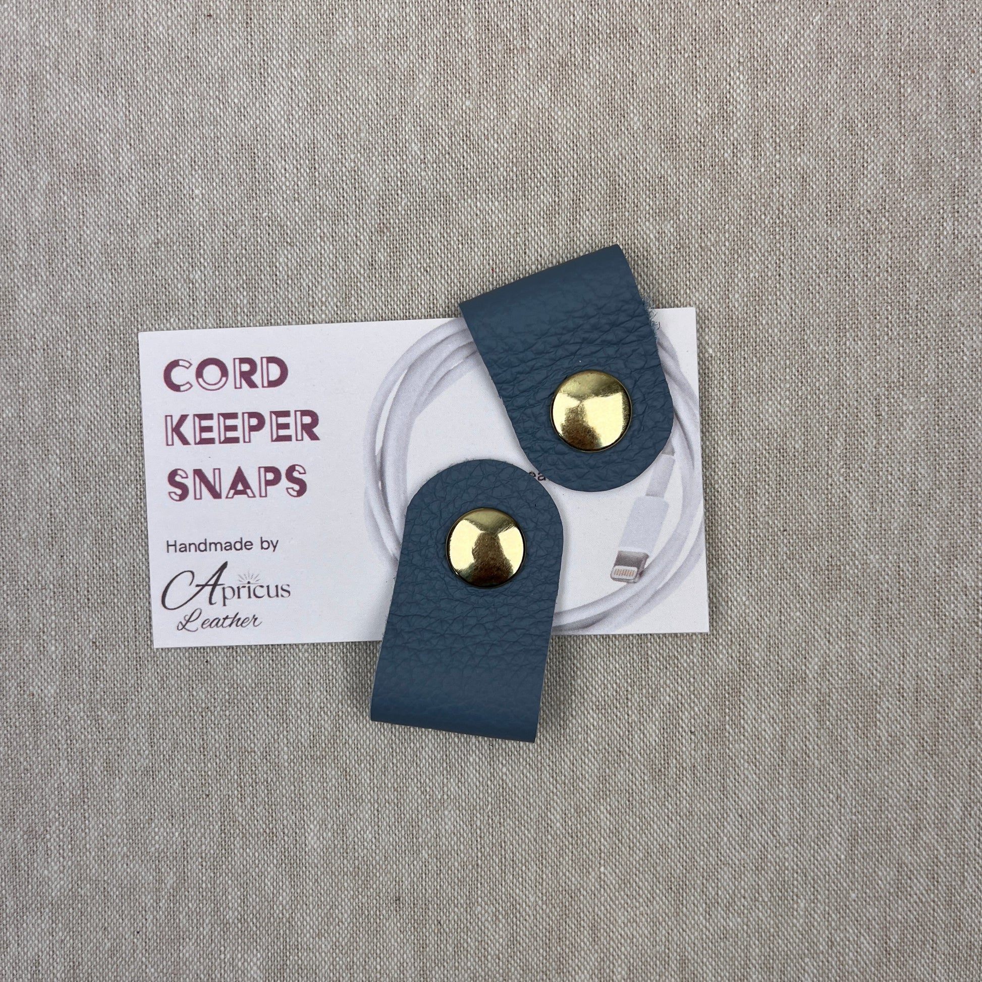 A pair of dark teal textured leather cord keepers with gold-tone snaps, shown with a white product card on a gray textured fabric background. The card reads 'CORD KEEPER SNAPS' and 'Handmade by Apricus Leather'.