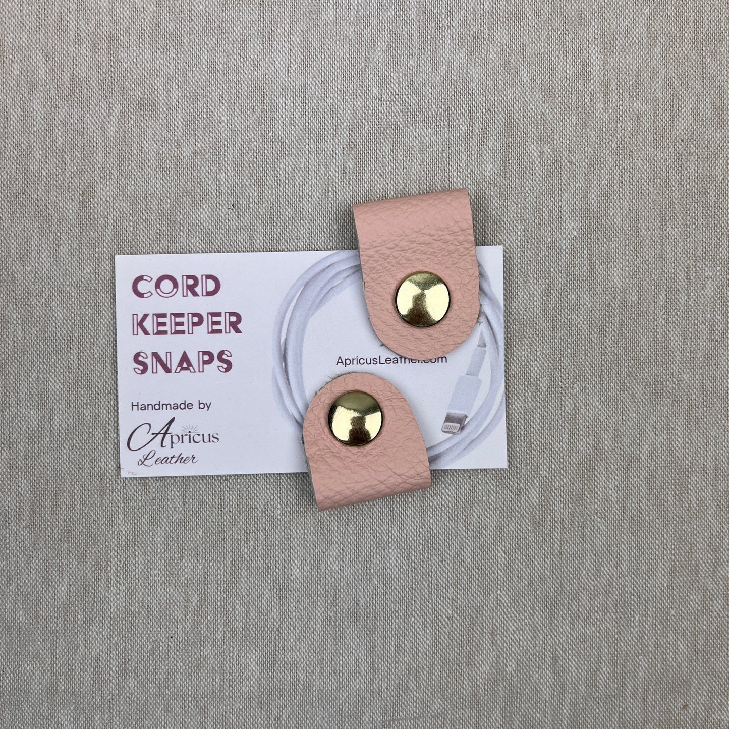 A pair of blush pink leather cord keepers with gold-tone snap buttons, shown with a white branded card on a textured gray fabric background.