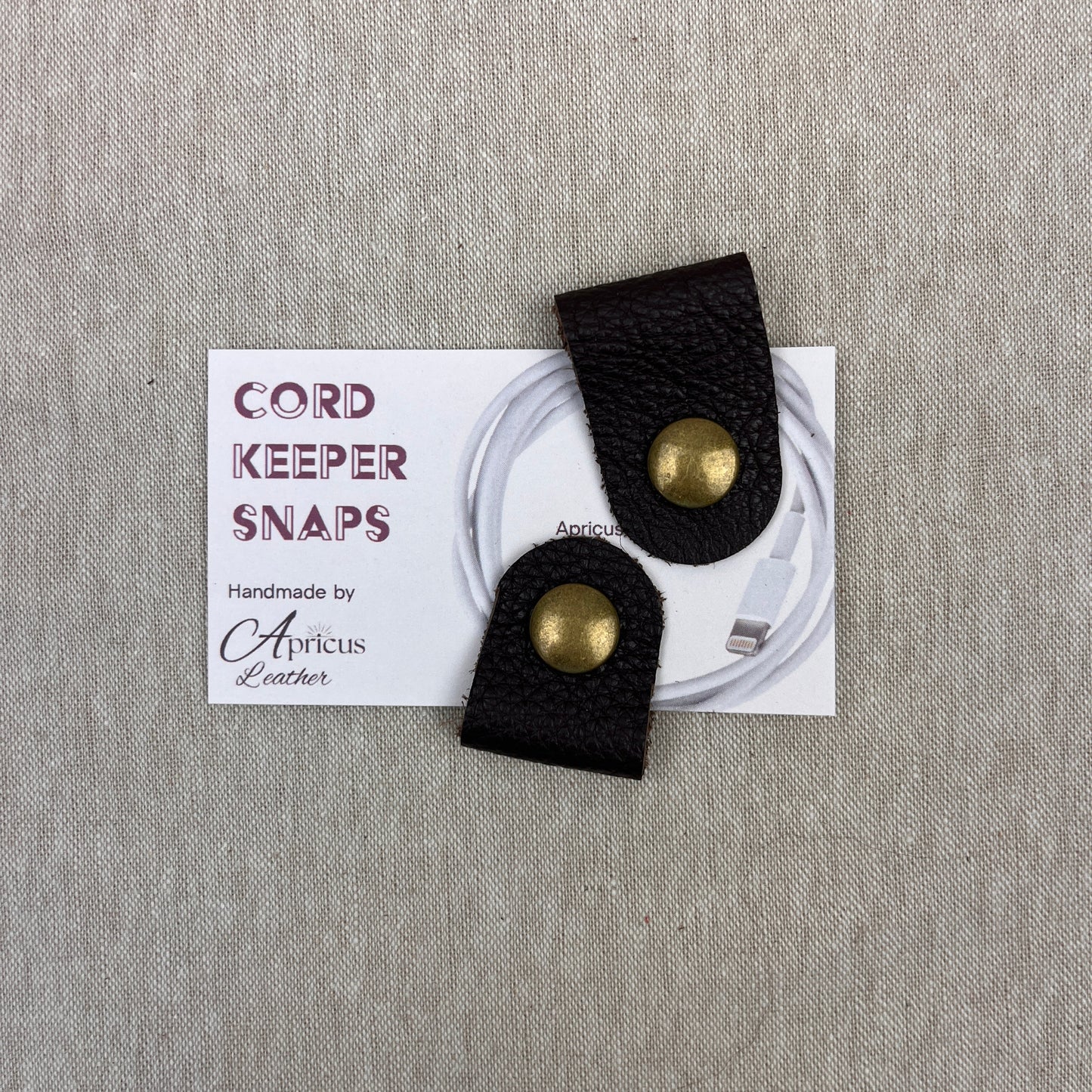 Two brown textured cord keeper snaps with antique brass-tone metal snaps, displayed on a white backing card over a gray fabric background.