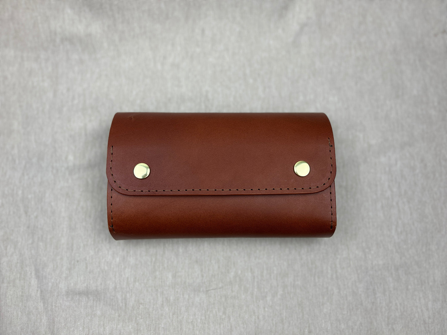 A brown leather cigar case with two gold-tone snap buttons, shown on a textured gray background.