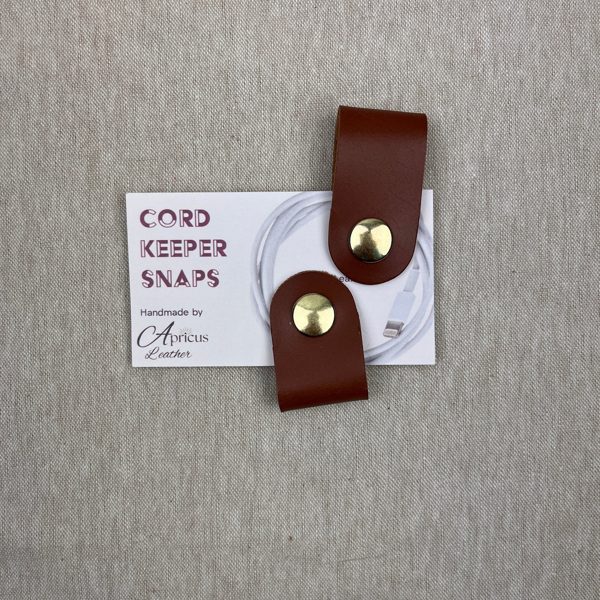 A pair of brown leather cord keeper snaps with gold-tone metal snaps, attached to a white paper tag that reads 'Cord Keeper Snaps' placed on a gray textured fabric background.