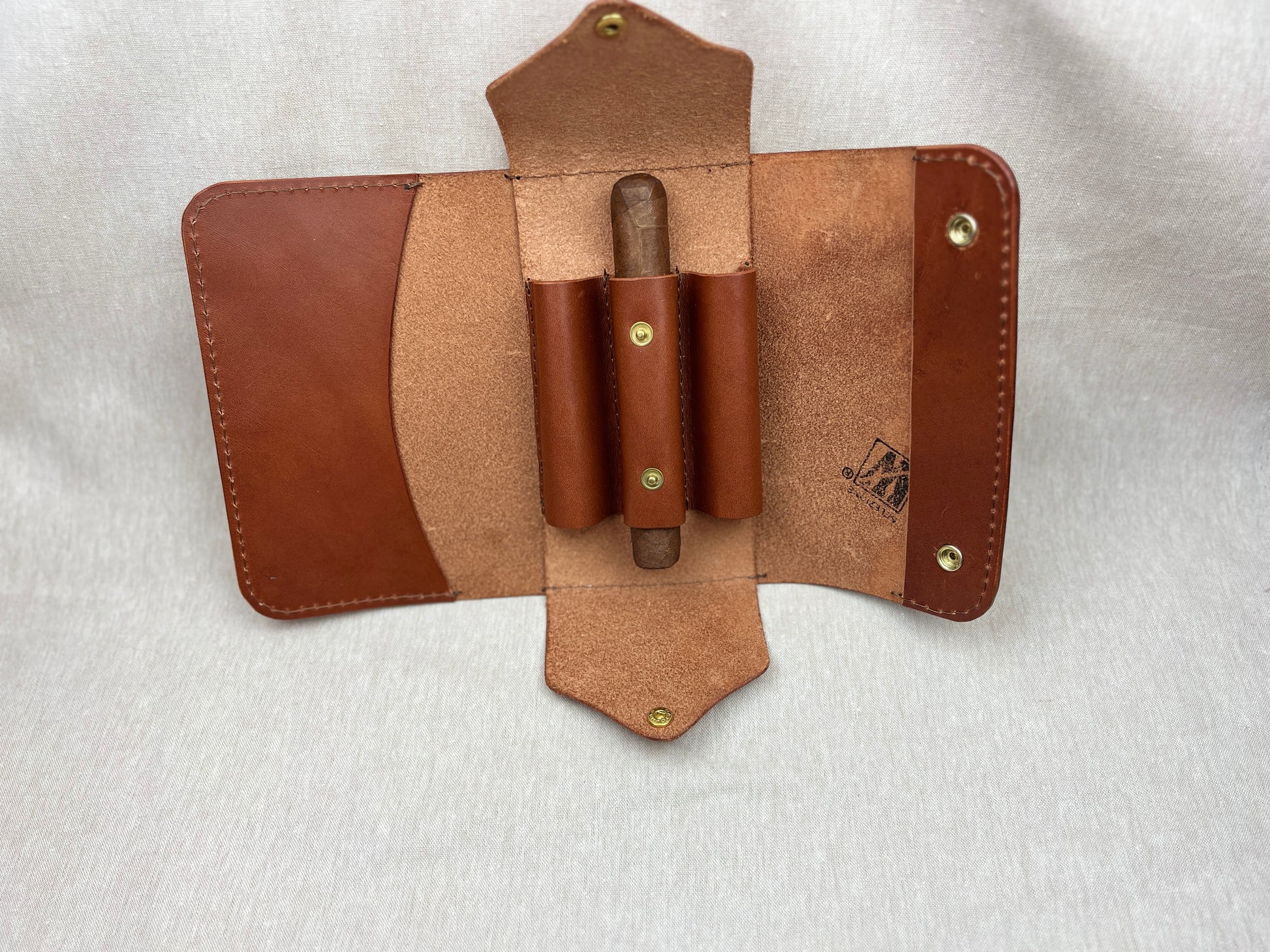 A brown leather cigar holder wallet shown open, featuring two vertical pouches holding cigars, a card pocket on the left, and a strap closure on the right, photographed on a light gray fabric background.