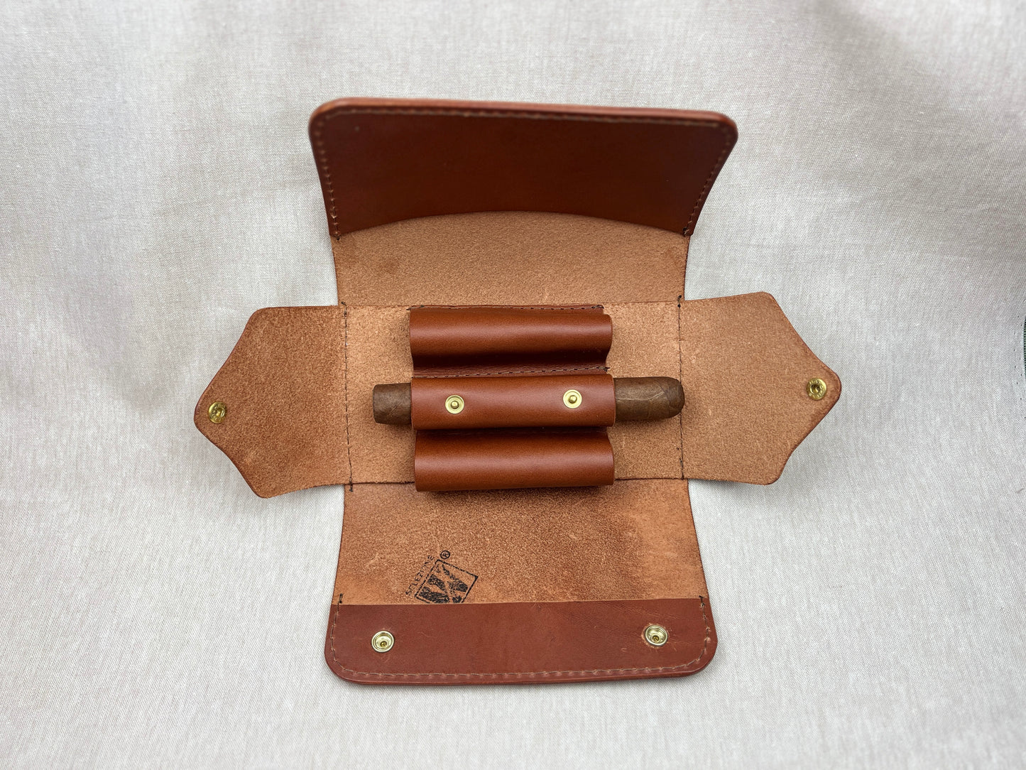 A brown leather cigar holder case shown open, featuring two rolled cigars in brown leather loops and brass snap fasteners, laid flat on a light gray textured background.