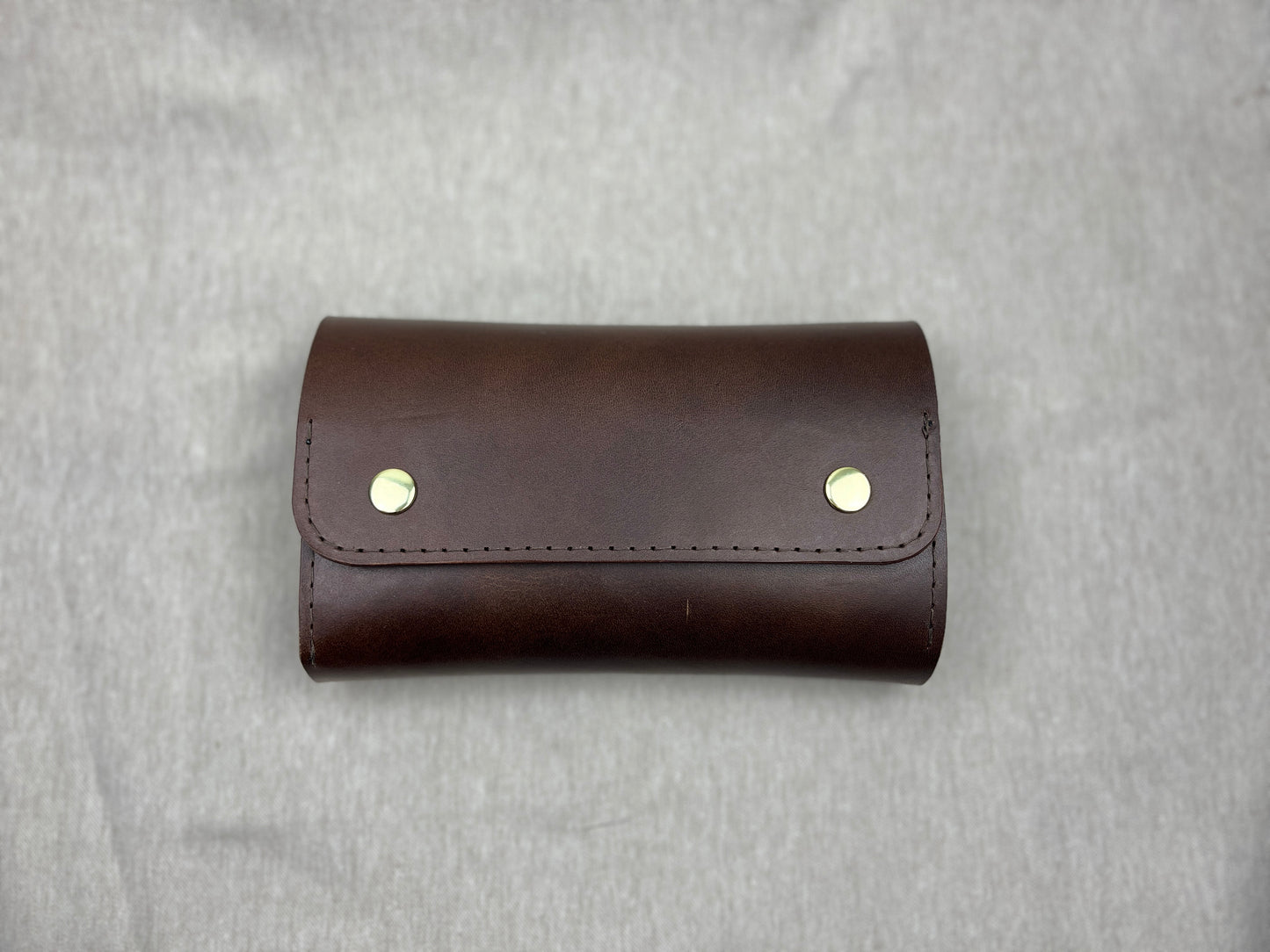 A dark brown leather cigar case with two gold-tone metal rivets on the front flap, photographed flat against a textured gray background.