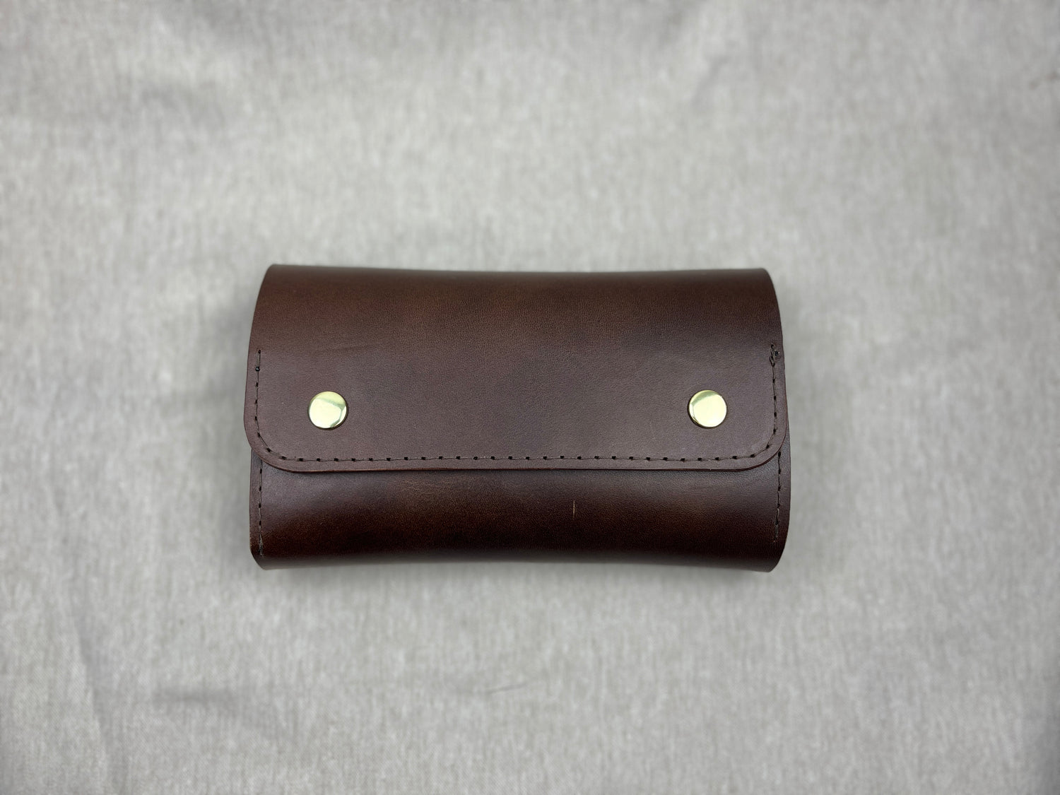 A dark brown leather cigar case with two gold-tone metal rivets on the front flap, photographed flat against a textured gray background.