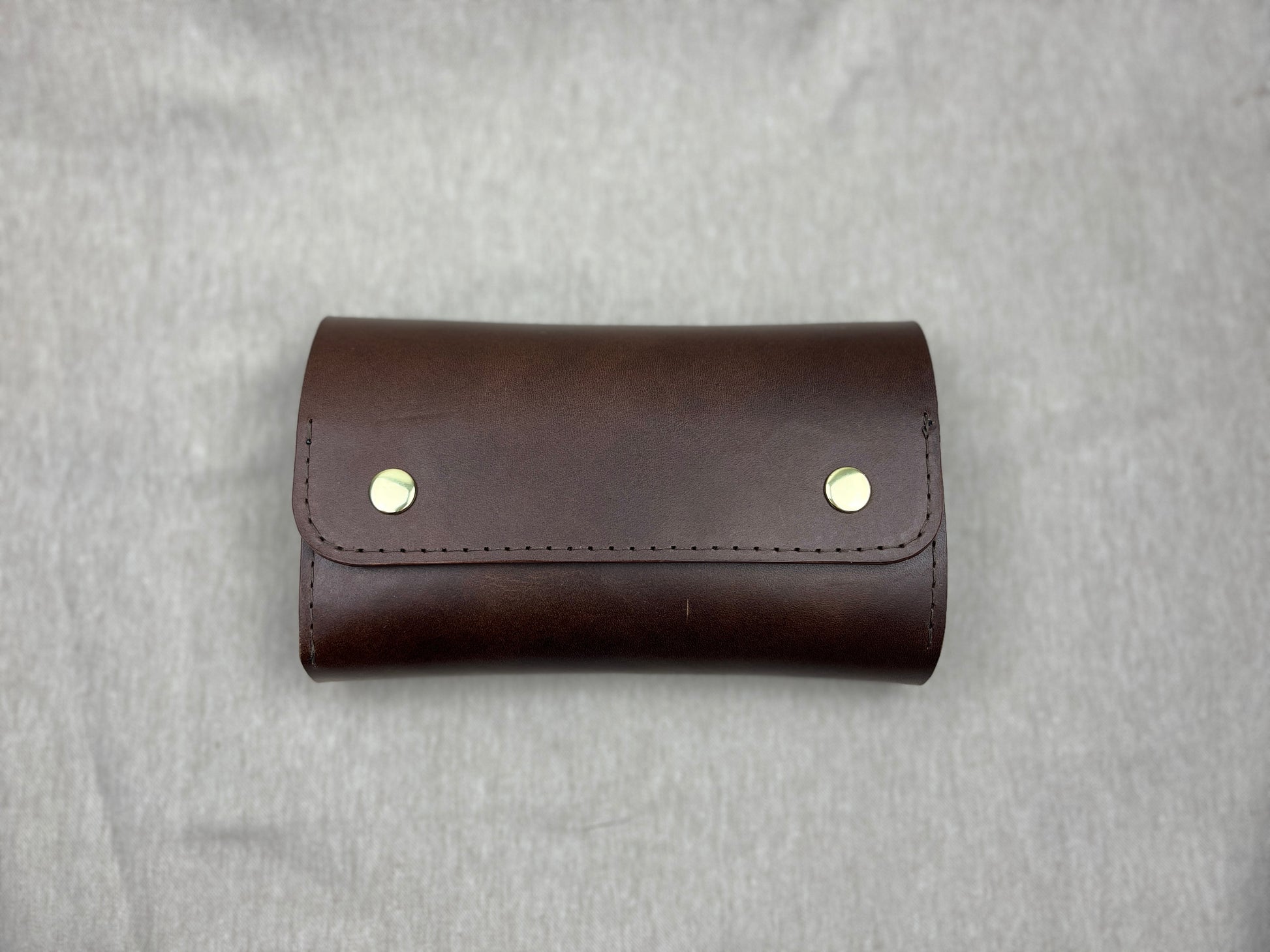 A dark brown leather cigar case with two gold-tone metal rivets on the front flap, photographed flat against a textured gray background.