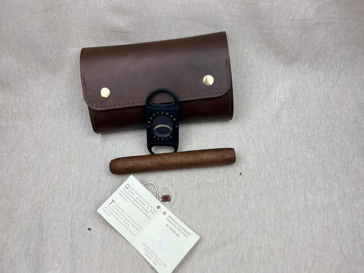 A brown leather cigar case with brass snap buttons, a black cigar cutter tool, a brown cigar, and a white paper tag, arranged on a light gray fabric background.