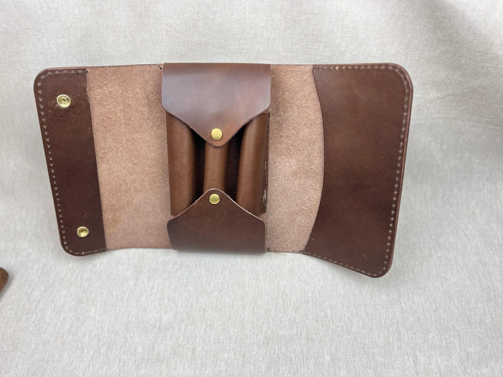 A brown leather cigar case with suede-like beige panels and brass snap buttons, laid flat on a light gray textured background.