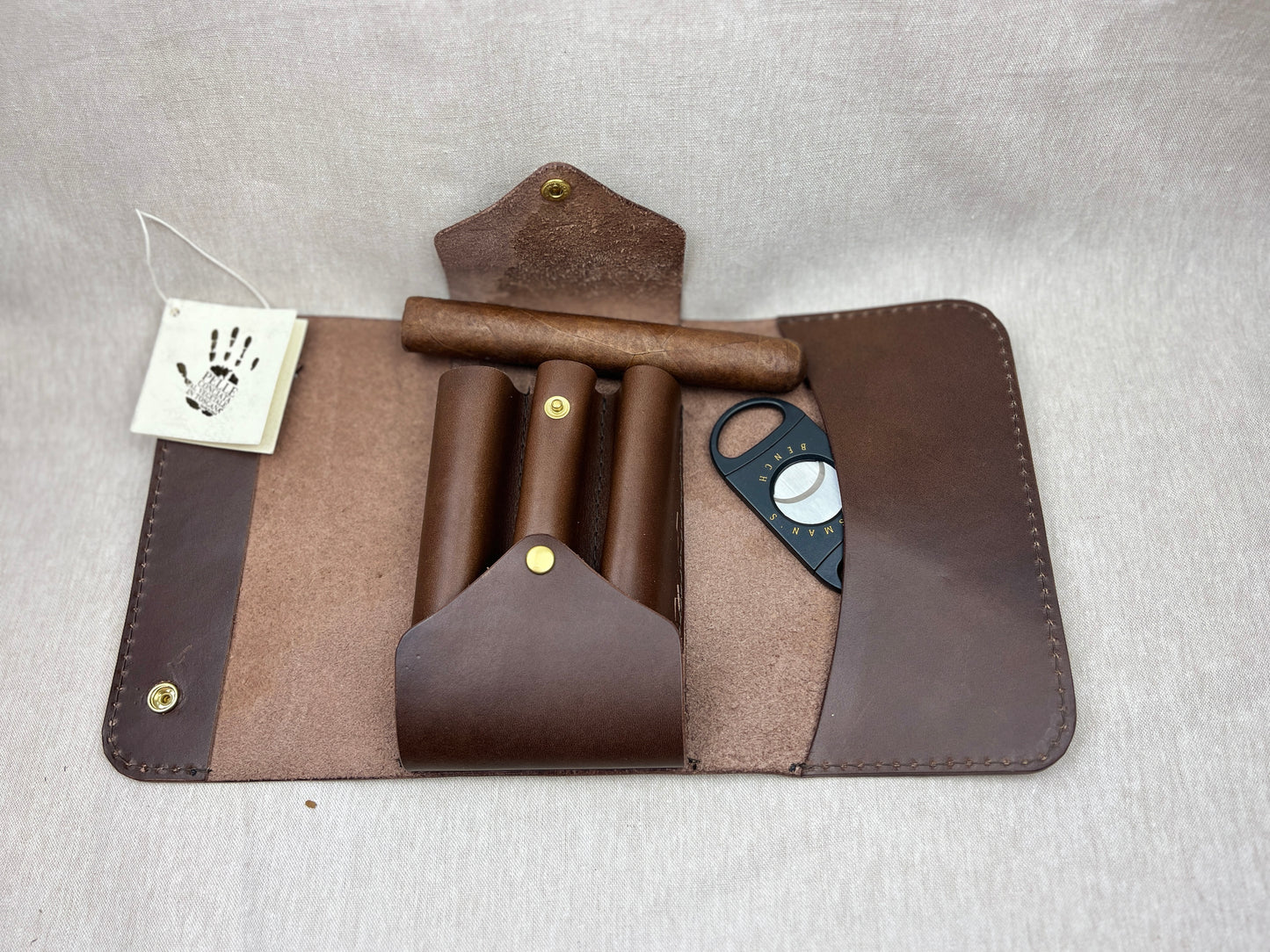 A brown leather cigar storage case opened flat on a light gray fabric background, containing a cigar, three rolled cigars in wooden holders, a black cigar cutter, and a small white tag with a handprint logo.