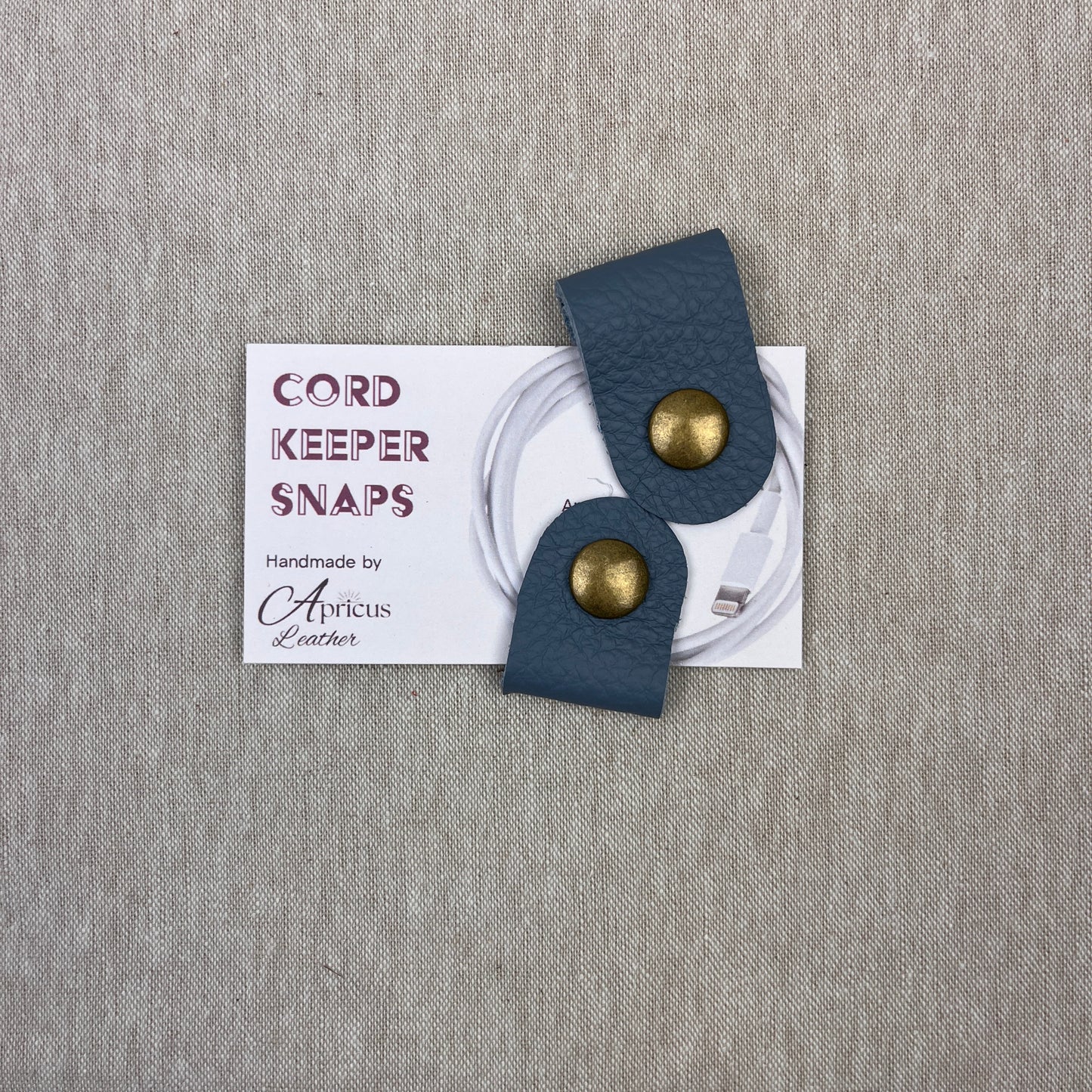 A pair of cornflower blue leather cord keepers with brass snaps, displayed on a white backing card that reads 'CORD KEEPER SNAPS' and 'Handmade by Apricus Leather', set against a textured beige background.