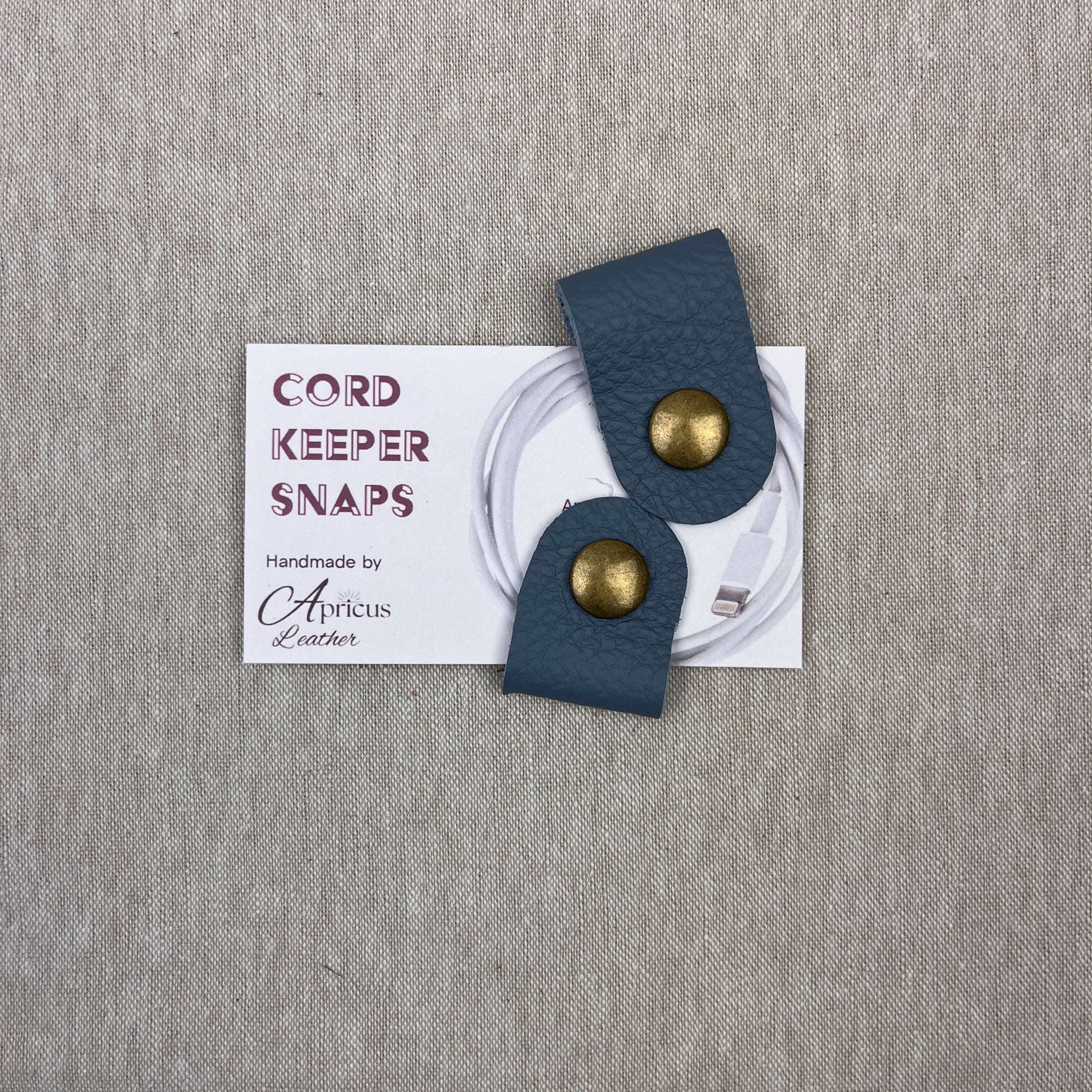 A pair of cornflower blue leather cord keepers with brass snaps, displayed on a white backing card that reads 'CORD KEEPER SNAPS' and 'Handmade by Apricus Leather', set against a textured beige background.