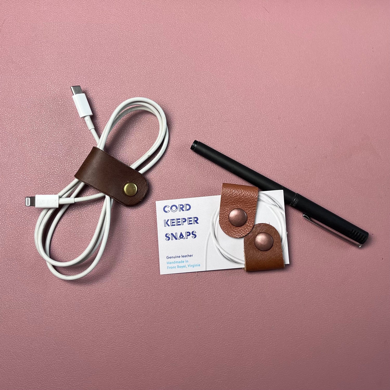 White cable with brown leather strap, black pen, and 'Cord Keeper Snaps' product on a pink background