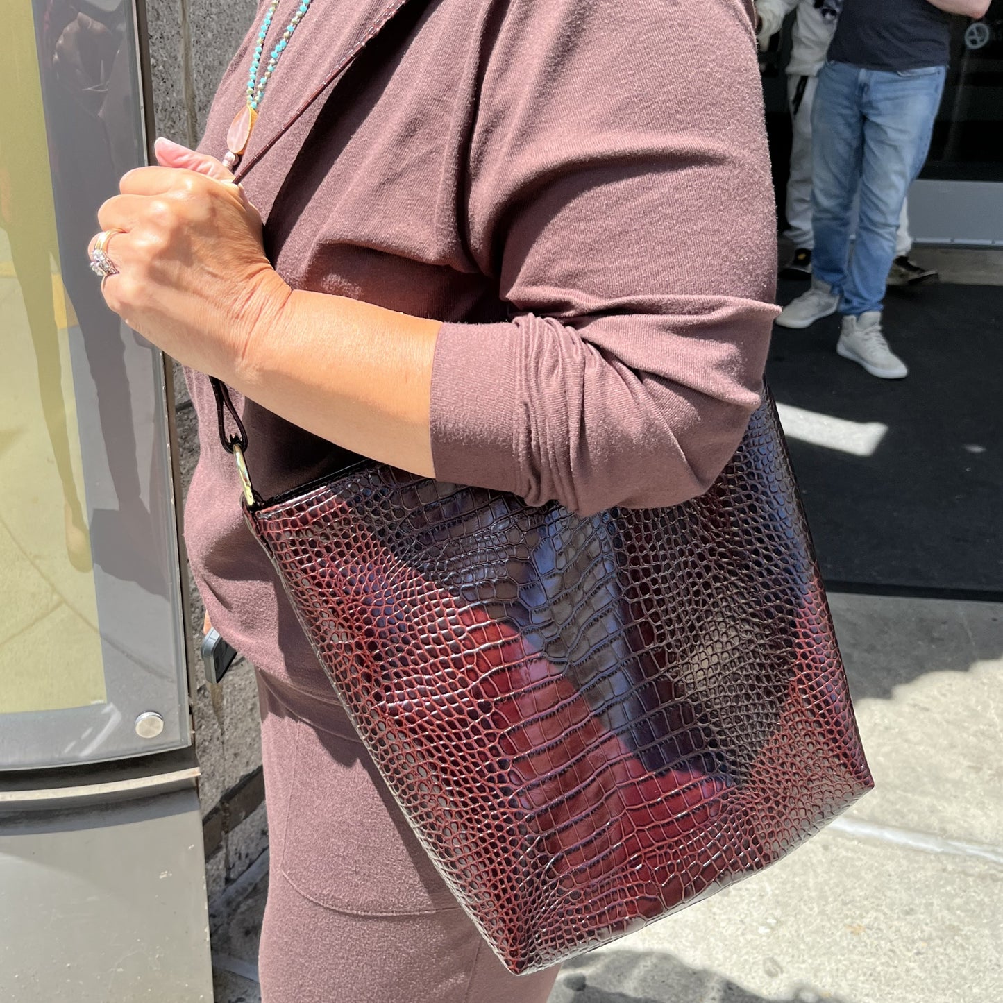 A person wearing a mauve-brown outfit carries a large rectangular shoulder bag with a glossy crocodile-embossed texture in a deep burgundy tone. The photo is taken outdoors near a storefront, with part of another person visible in the background.