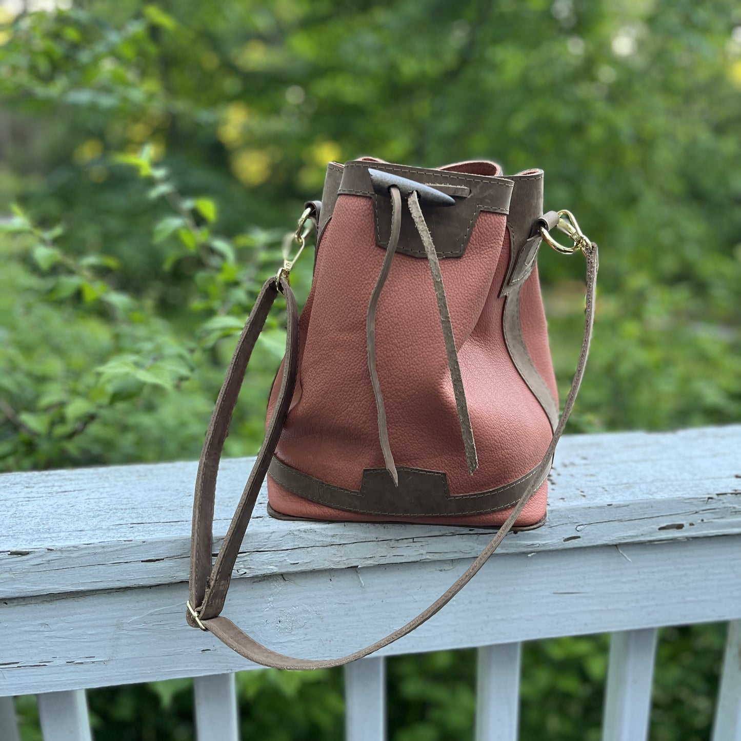 Apricot leather bucket bag with brown leather trim and a matching shoulder strap, resting on a white wooden railing with a blurred green garden background.