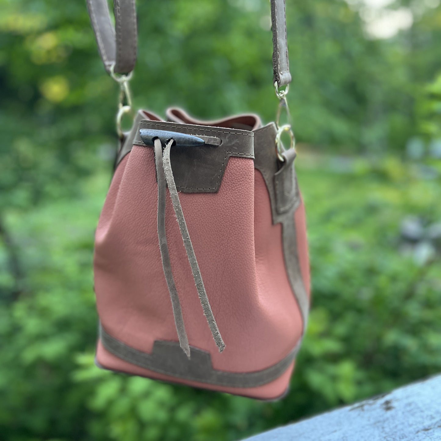 An apricot color leather bucket bag with brown leather trim and drawstring details, hanging outdoors against a blurred green foliage background.