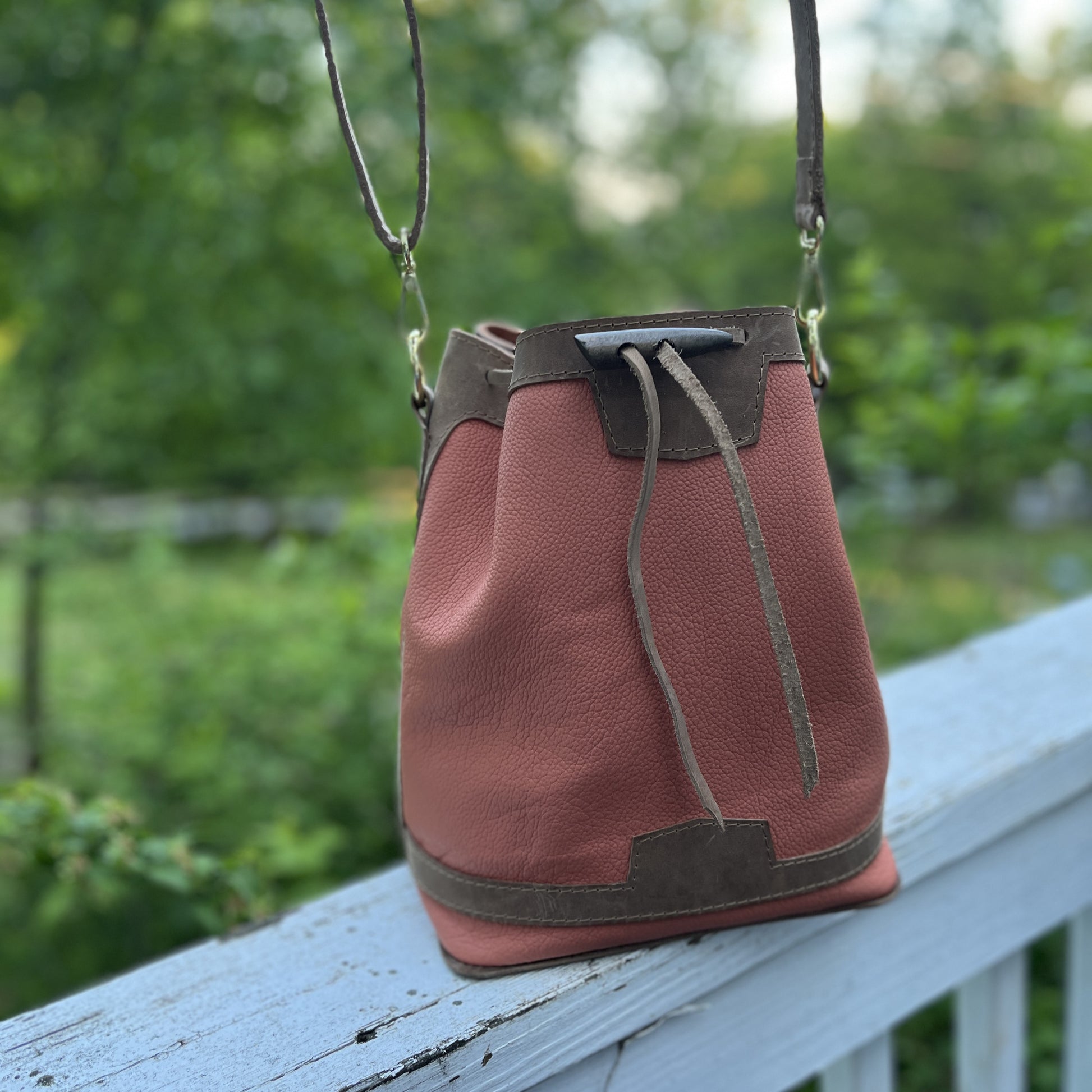 An apricot leather bucket bag with dark brown trim and drawstring details, resting on a weathered blue-gray wooden railing with a blurred green garden background.
