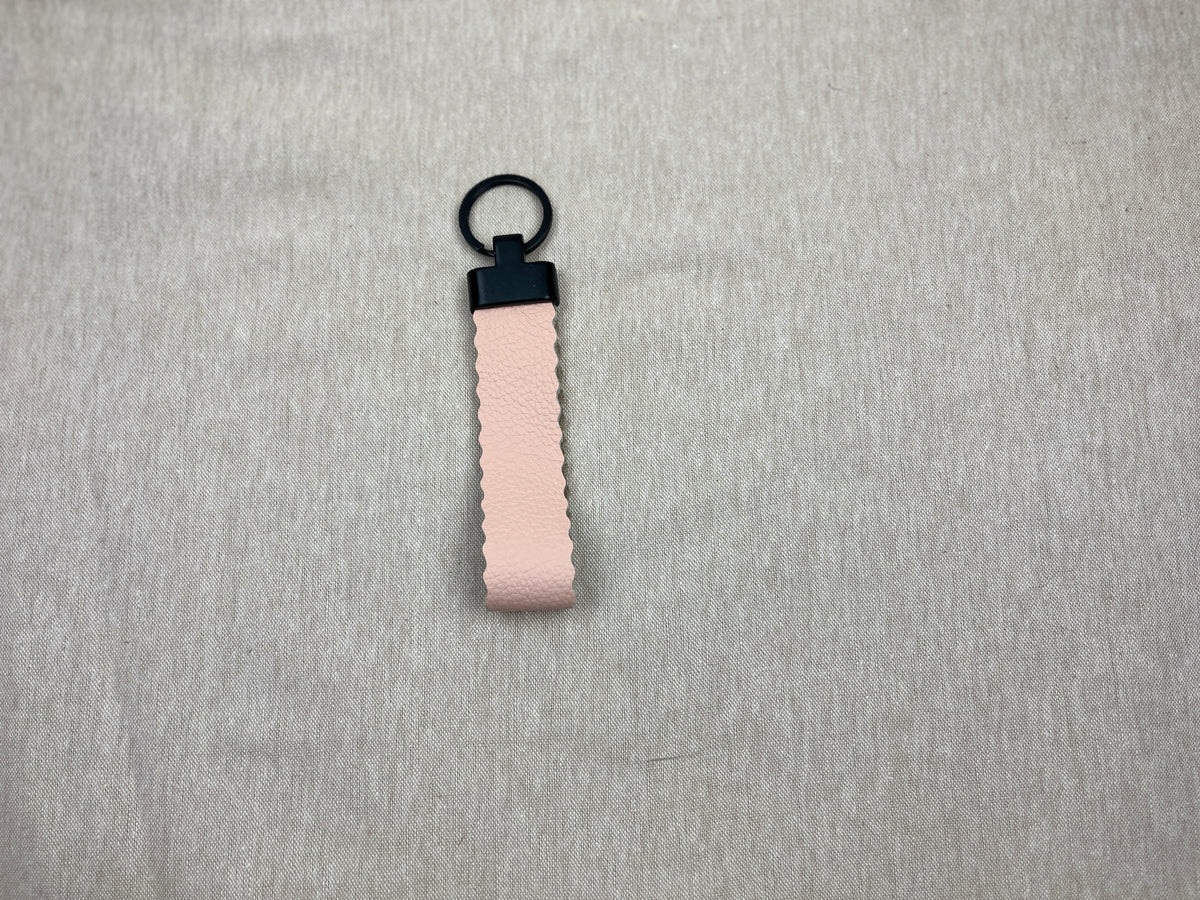 A small light pink textured leather keychain loop with a black plastic connector and black metal key ring, resting on a soft beige fabric background.