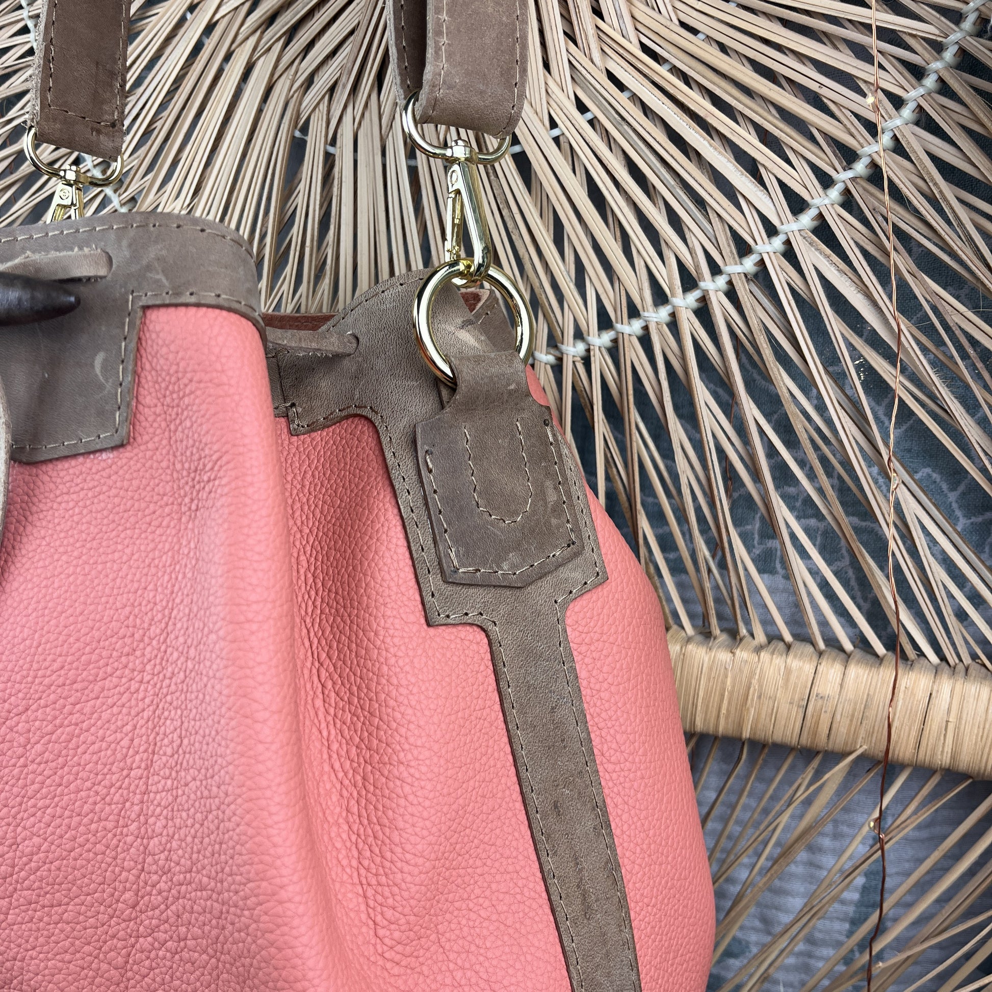 A close-up of a pink pebbled leather handbag with brown suede-like trim and strap details, hanging against a woven straw or palm leaf background.