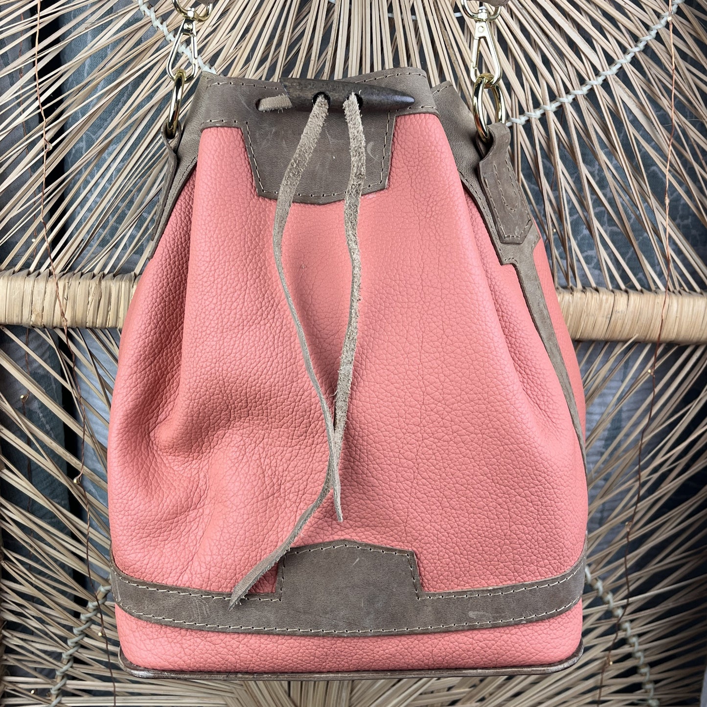 A dusty pink leather bucket bag with dark gray suede-like trim and drawstring closure, resting on a woven rattan chair.