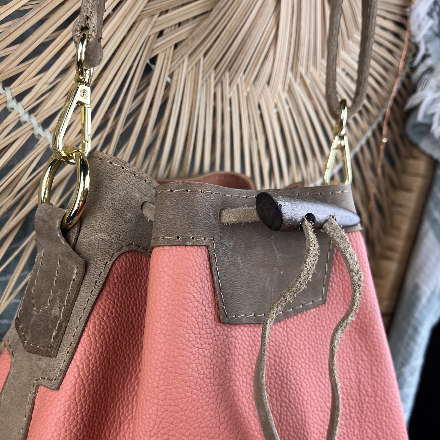 A close-up of a pink pebbled leather bucket bag with taupe suede trim, gold-tone hardware, and drawstring details, shown against a woven palm leaf background.