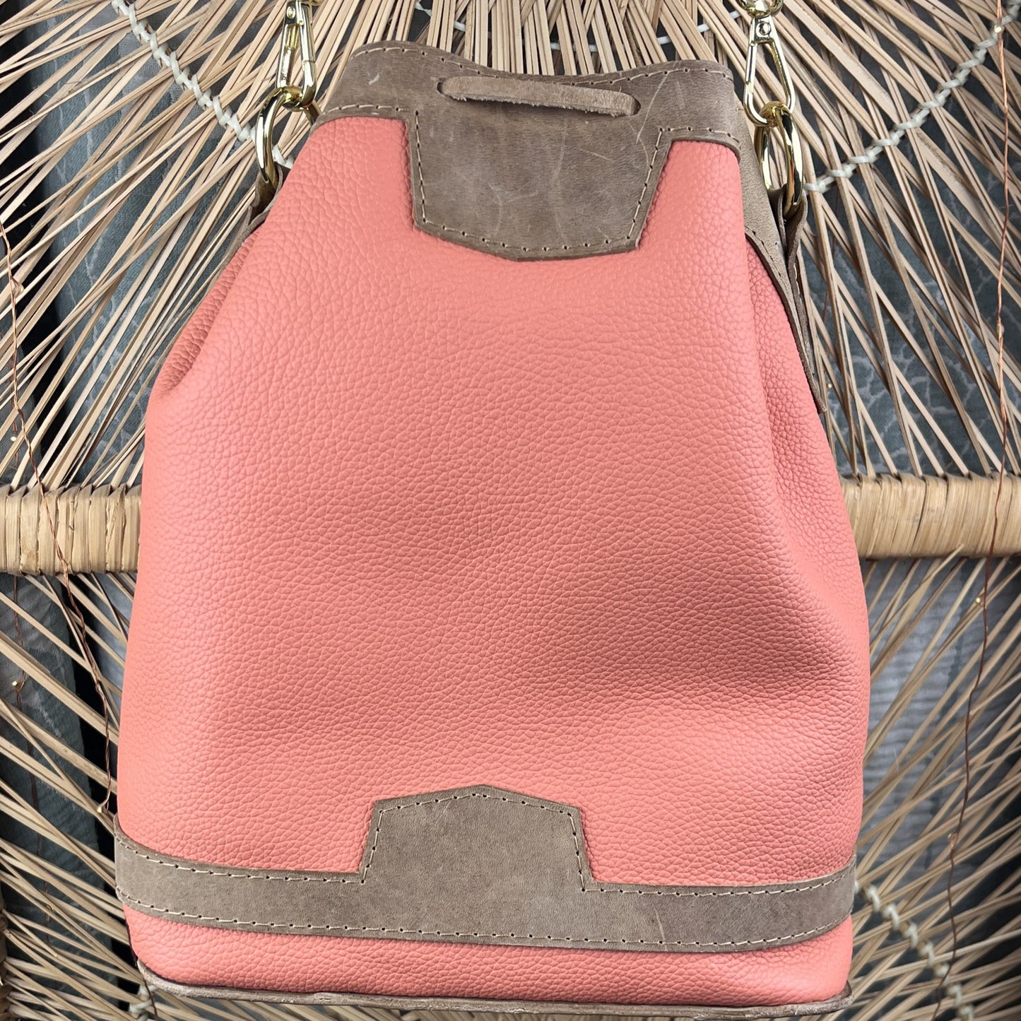 A pink pebbled leather bucket bag with taupe suede-like trim, shown resting on a woven rattan chair.