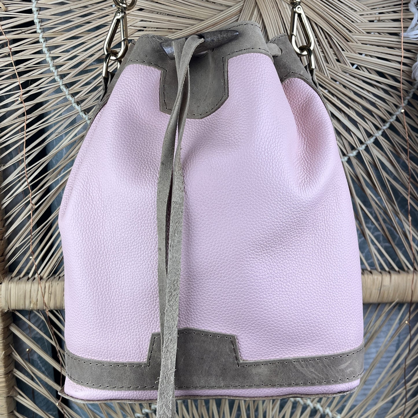 A light pink pebbled leather bucket bag with light brown trim and drawstring closure, shown hanging against a woven rattan background.