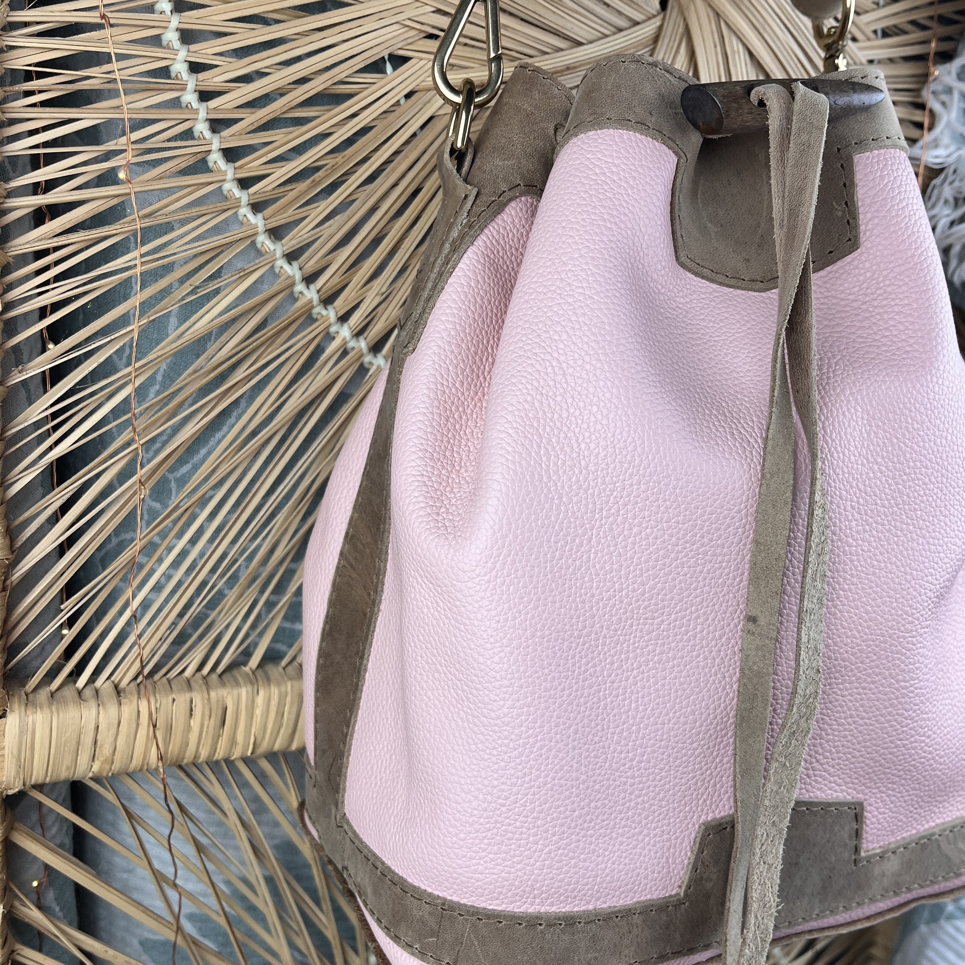 A light pink pebbled leather bucket bag with taupe suede trim and drawstring closure, shown hanging against a woven wicker background.
