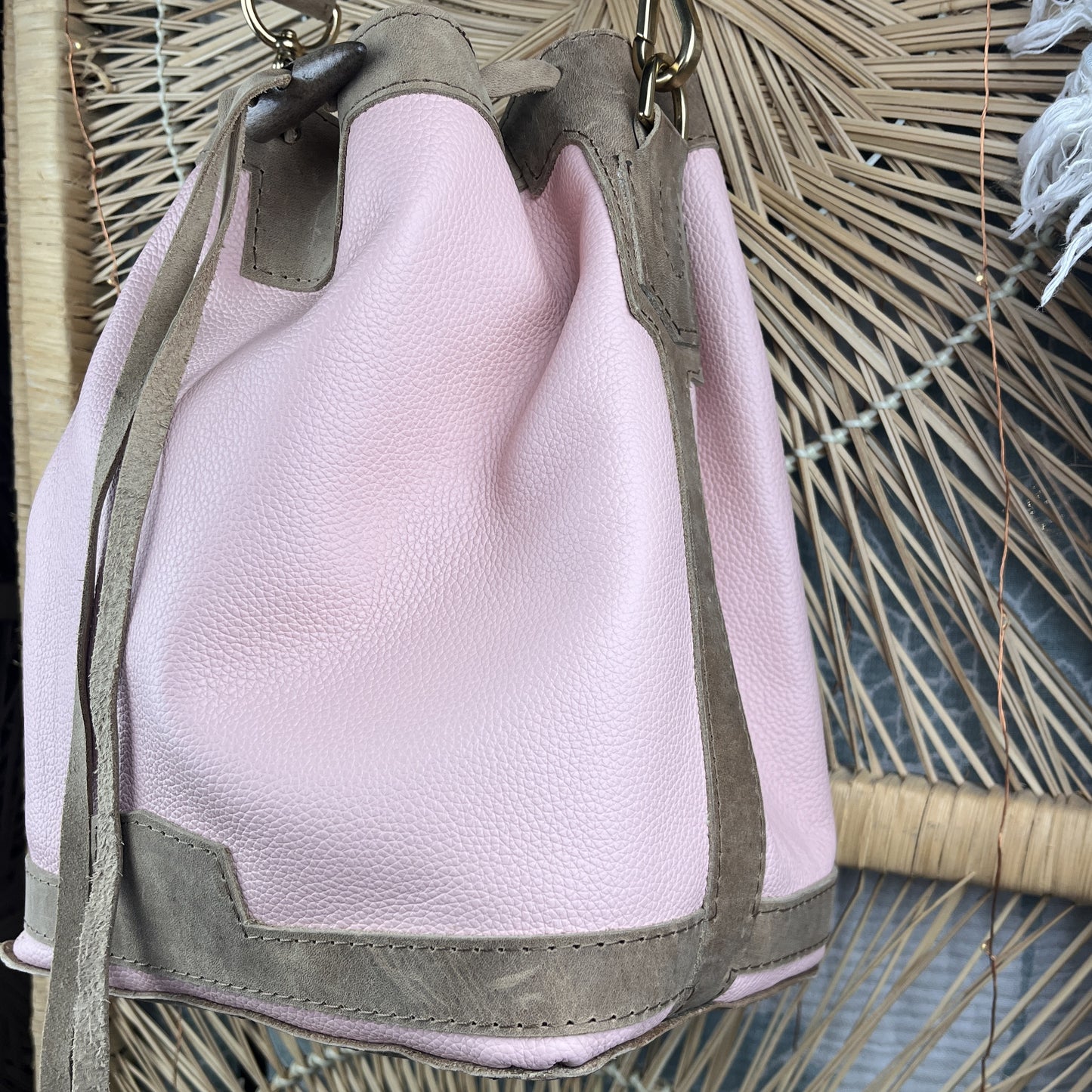 A light pink pebbled leather bucket bag with taupe suede trim and a matching shoulder strap, shown against a woven palm leaf background.