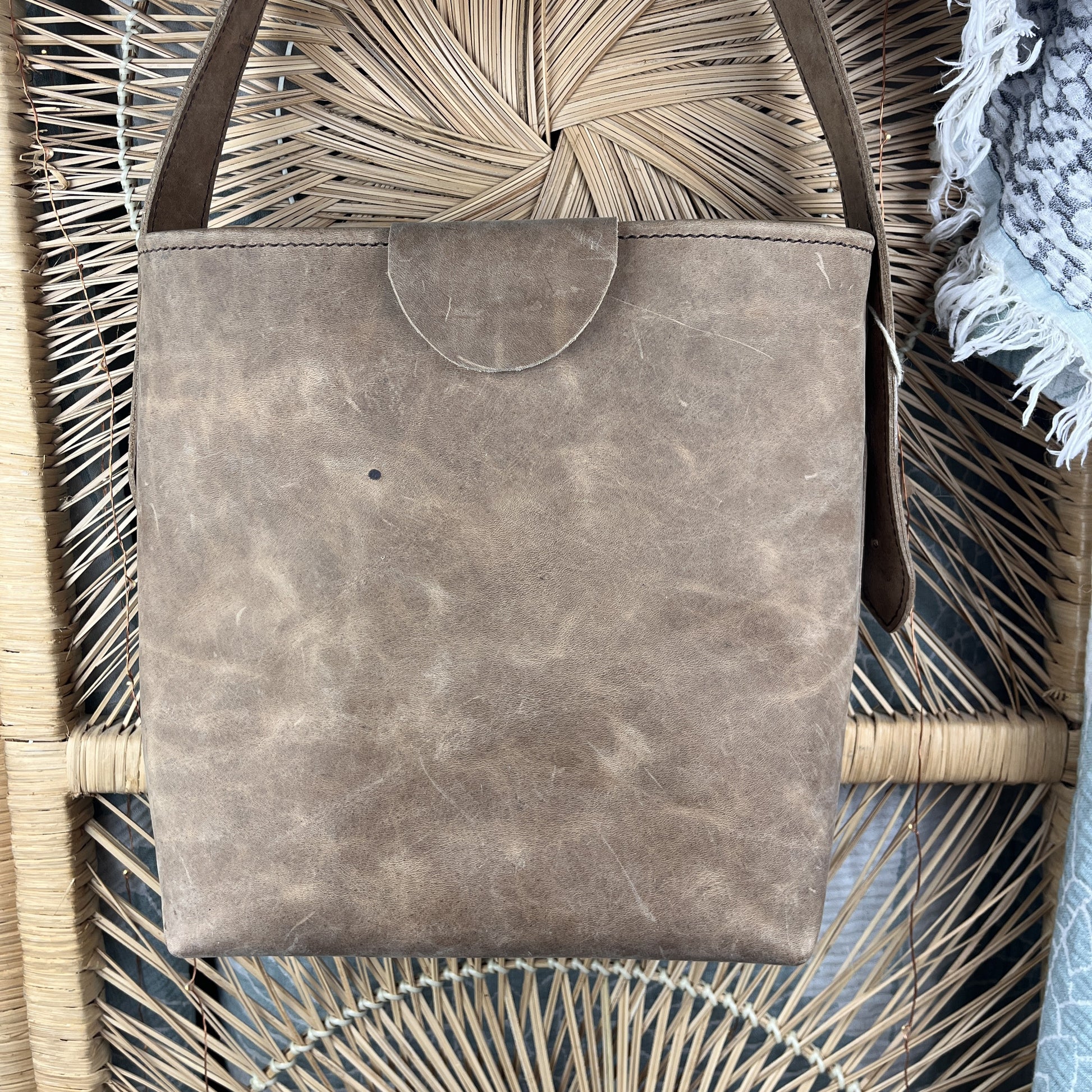 A distressed brown leather tote bag with a curved top flap, resting on a woven rattan chair.