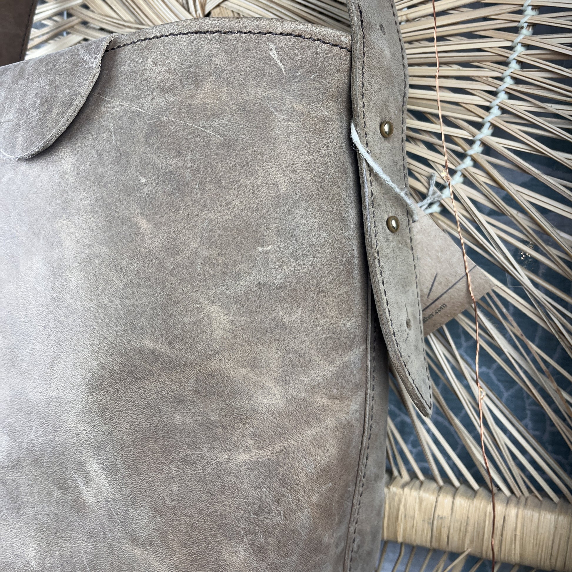 Close-up of a distressed gray leather bag strap with brass rivets, resting against a woven straw chair.