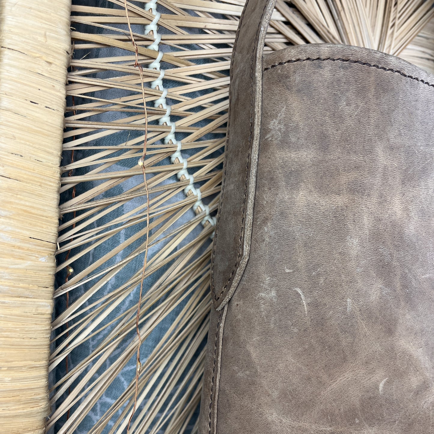 A close-up of a taupe-brown leather bag or pouch positioned next to a woven straw mat and a blue-gray textured surface.