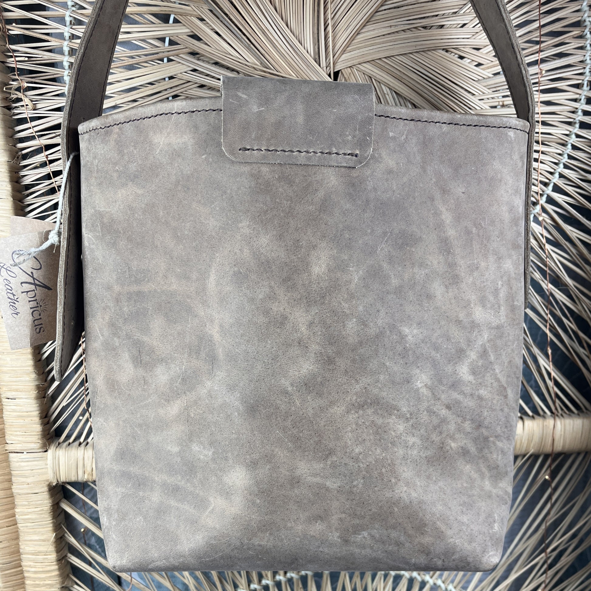 A distressed gray leather tote bag with dark gray straps, displayed on a woven straw chair.