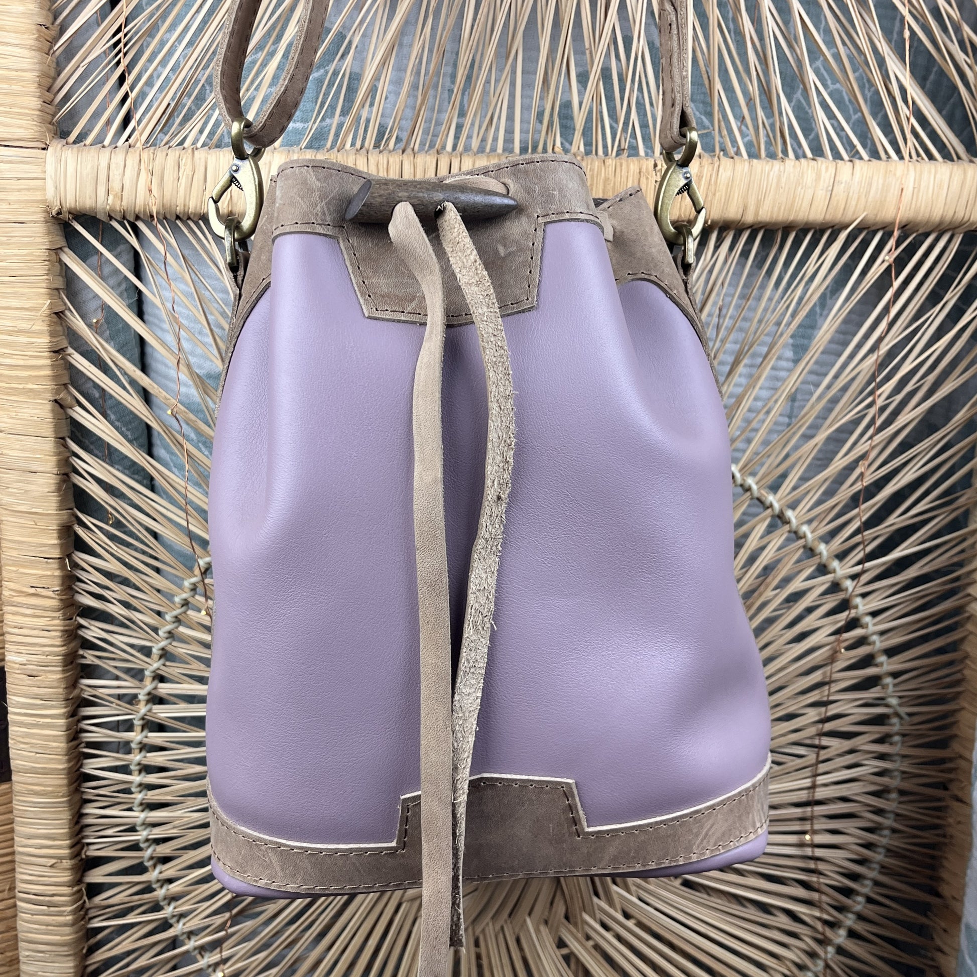A wisteria purple leather bucket bag with taupe trim and drawstring closure, displayed against a woven rattan chair backdrop.