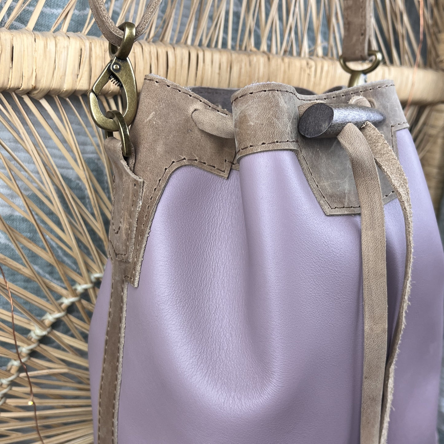 A close-up of a drawstring bucket bag in light lavender leather with tan suede trim and brass-tone hardware, shown against a woven straw background.