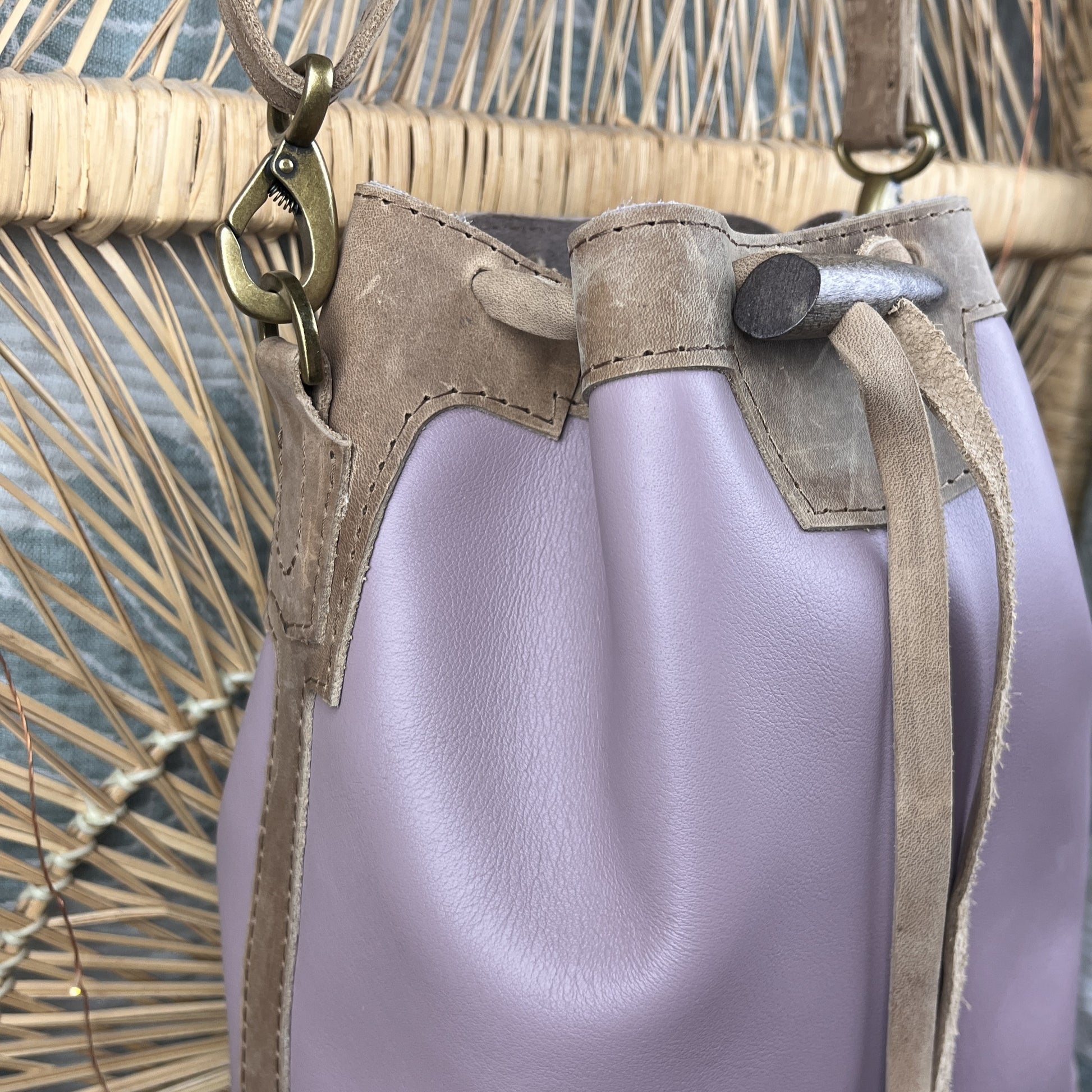 A close-up of a drawstring bucket bag in light lavender leather with tan suede trim and brass-tone hardware, shown against a woven straw background.