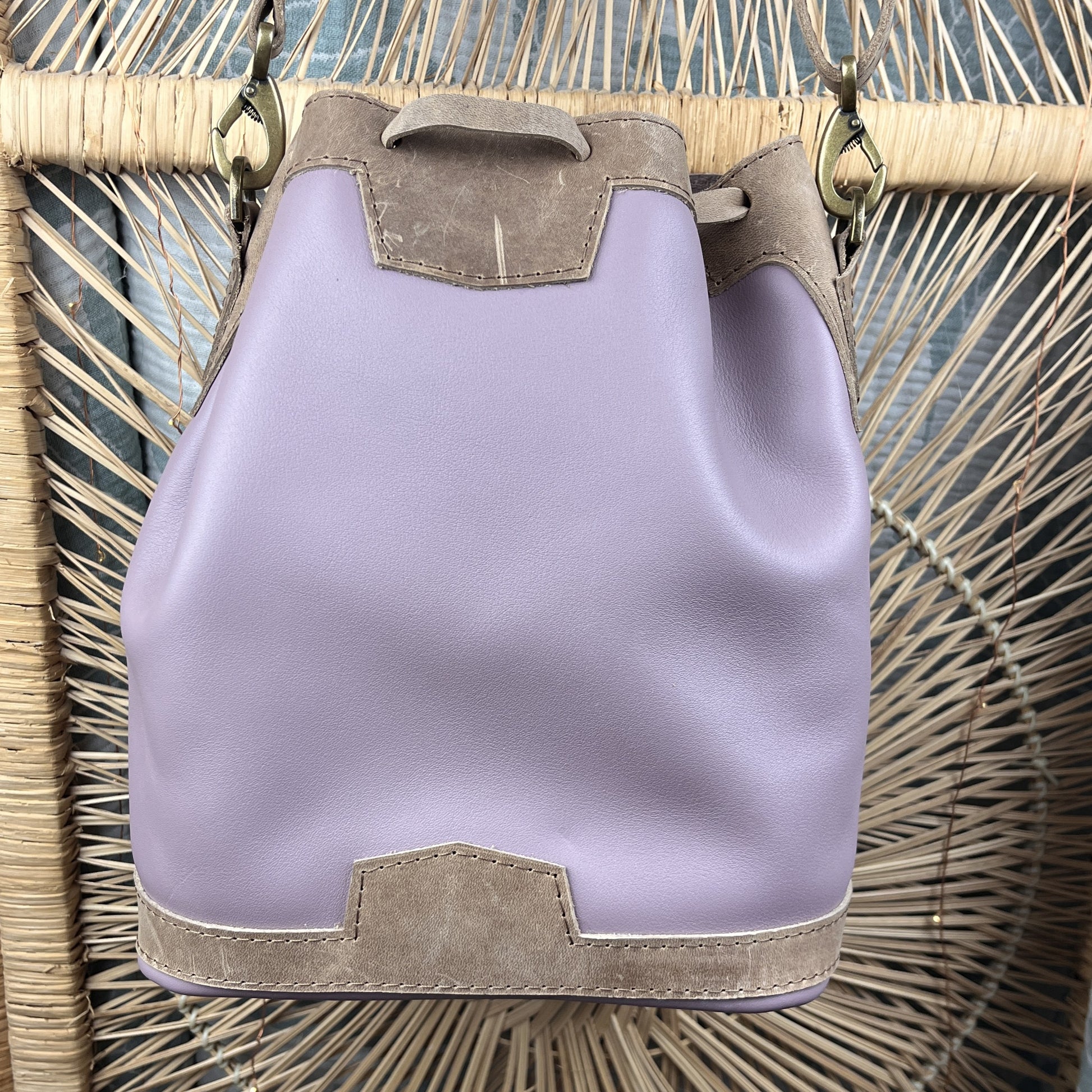 A lavender leather bucket bag with taupe suede trim and gold-tone hardware, hanging against a woven rattan backdrop.