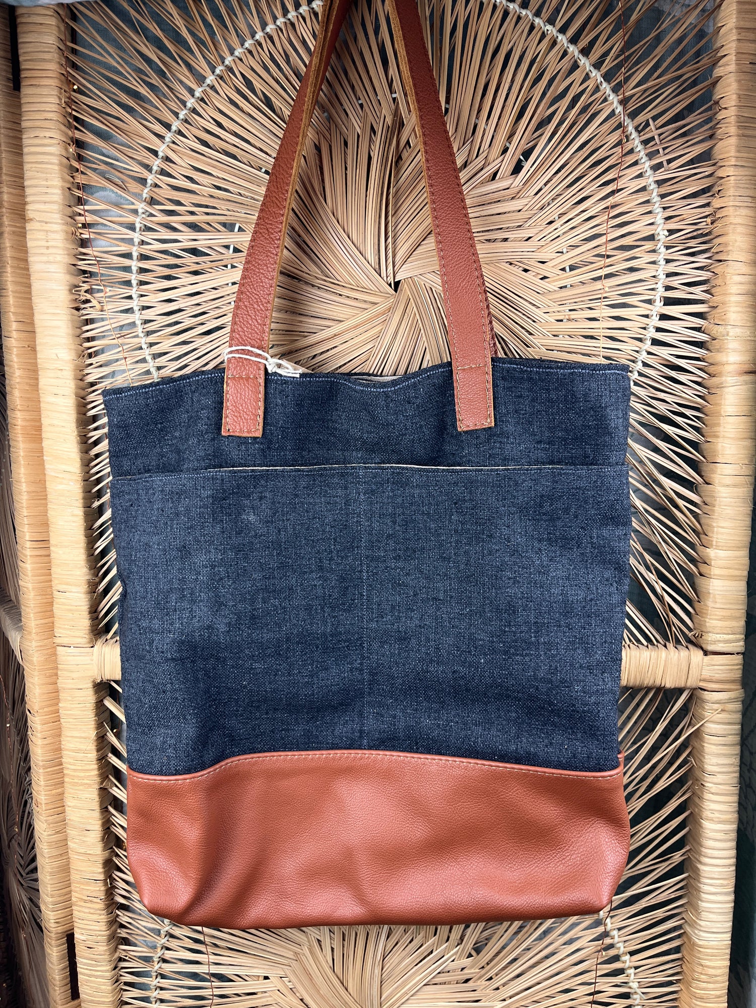 A handmade tote bag made of dark blue denim and brown leather, featuring two brown shoulder straps, laid flat on a woven straw mat.