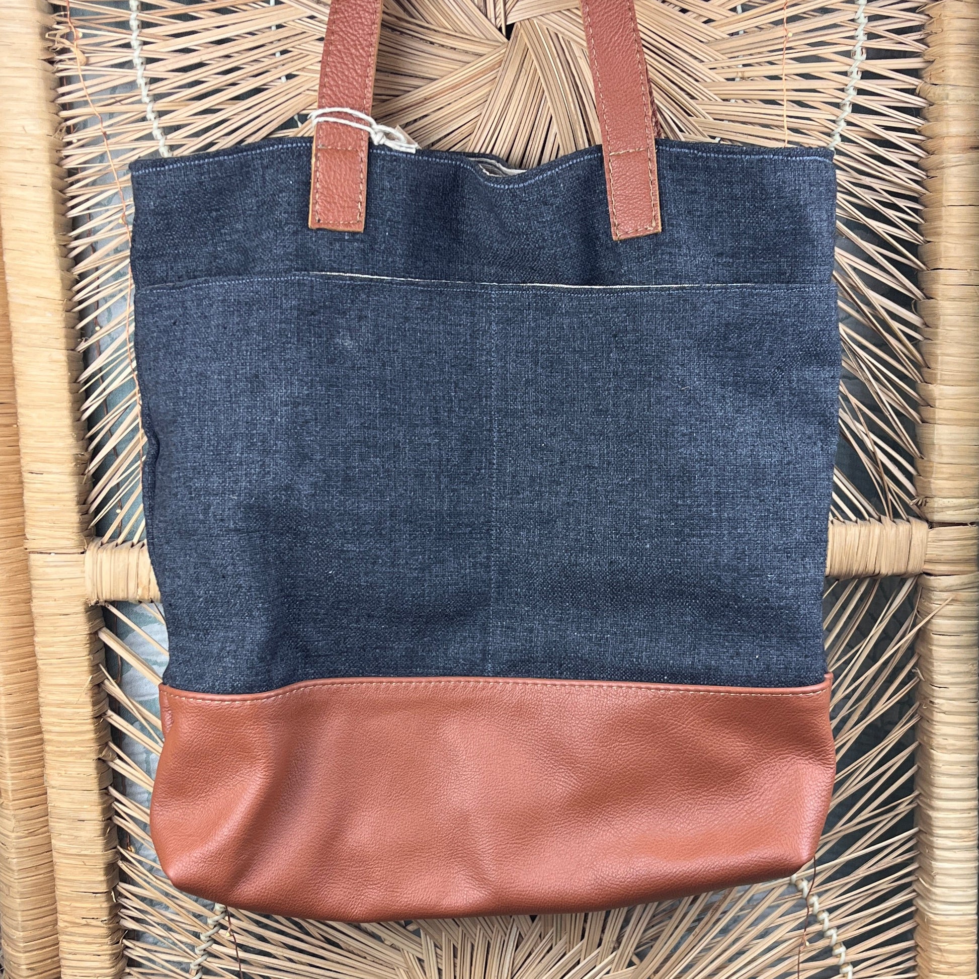 A tote bag with a dark gray linen-textured body, a brown leather side panel, and brown leather shoulder straps, displayed on a woven rattan surface.