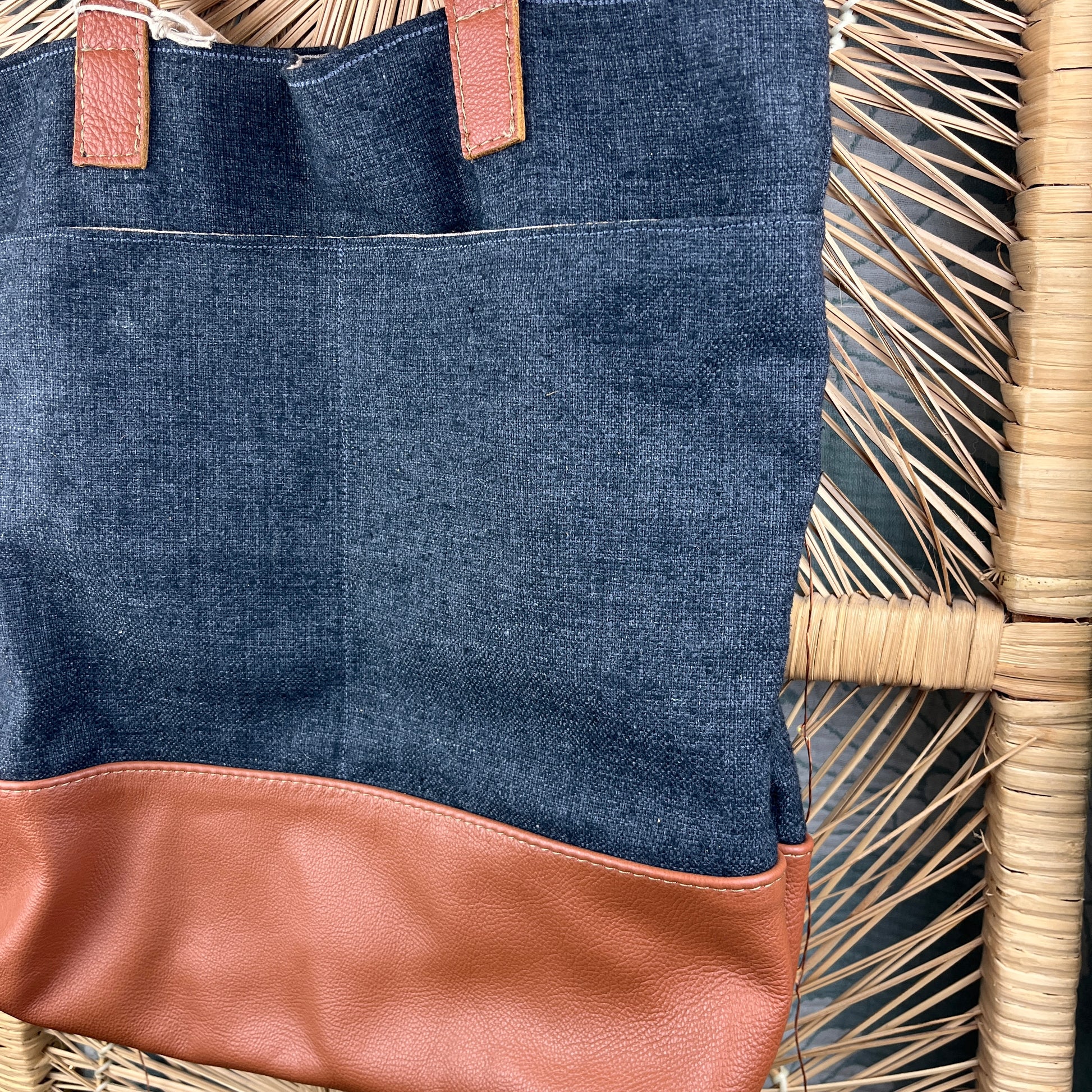 A close-up of a tote bag made of dark blue denim with a tan leather bottom panel and matching leather handle tabs, resting on a woven rattan chair.