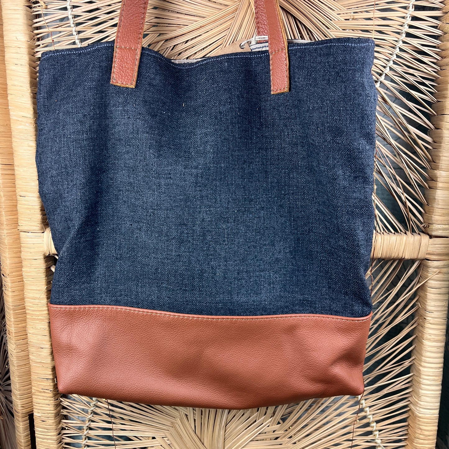A dark blue denim tote bag with a tan faux-leather bottom panel and matching handles, displayed on a woven wicker chair.