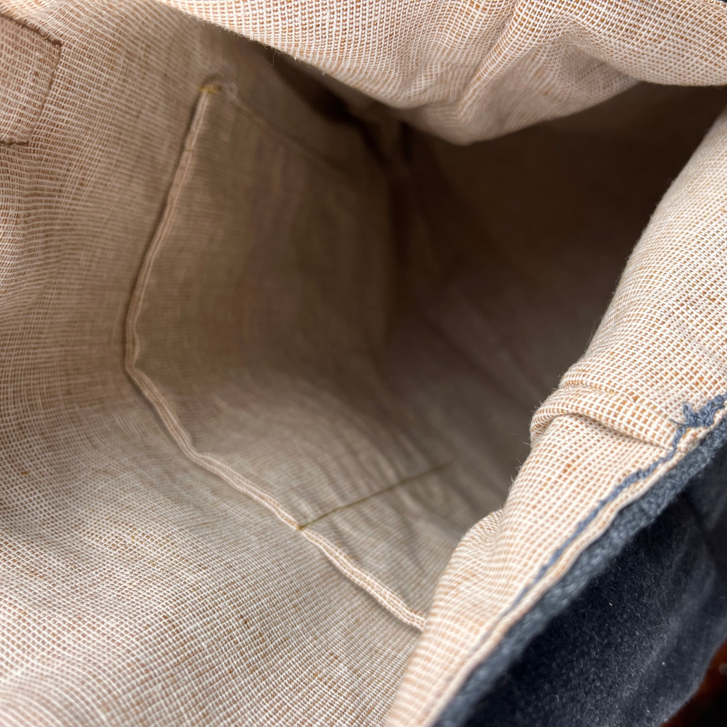 Close-up of a beige woven fabric bag or pouch showing the interior lining, a stitched pocket, and contrast blue stitching along the edge.