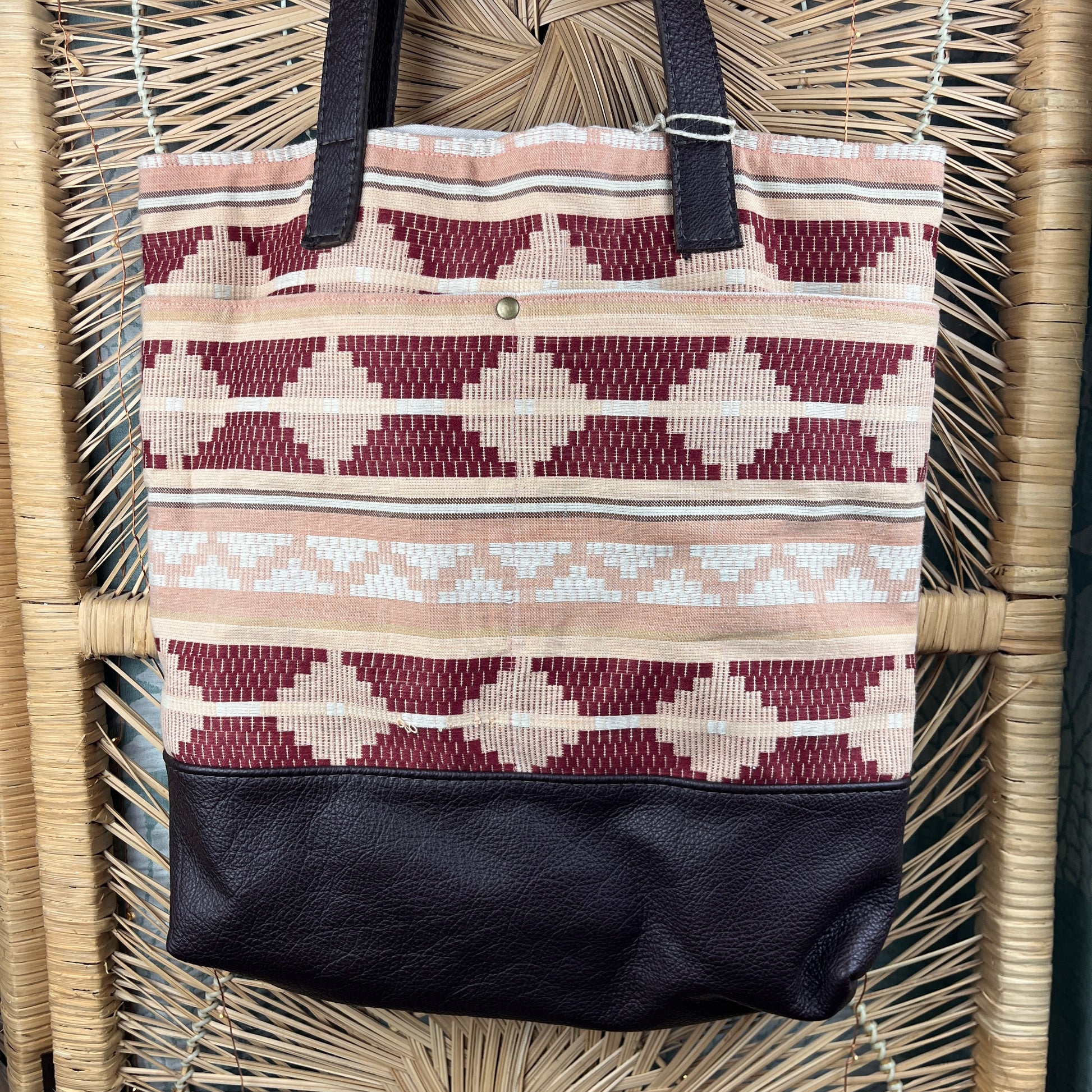 A woven tote bag with a geometric pattern in beige, cream, and burgundy, featuring a black faux-leather bottom and black shoulder straps, hanging on a wicker chair.