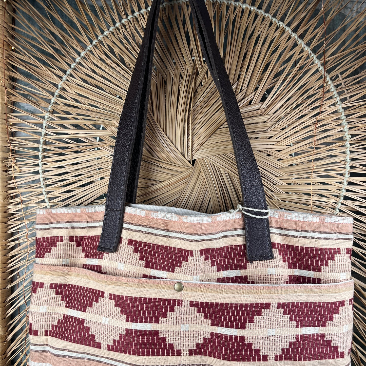A woven tote bag with a geometric diamond pattern in burgundy, beige, and white, featuring dark brown faux-leather shoulder straps, displayed against a circular woven straw background.