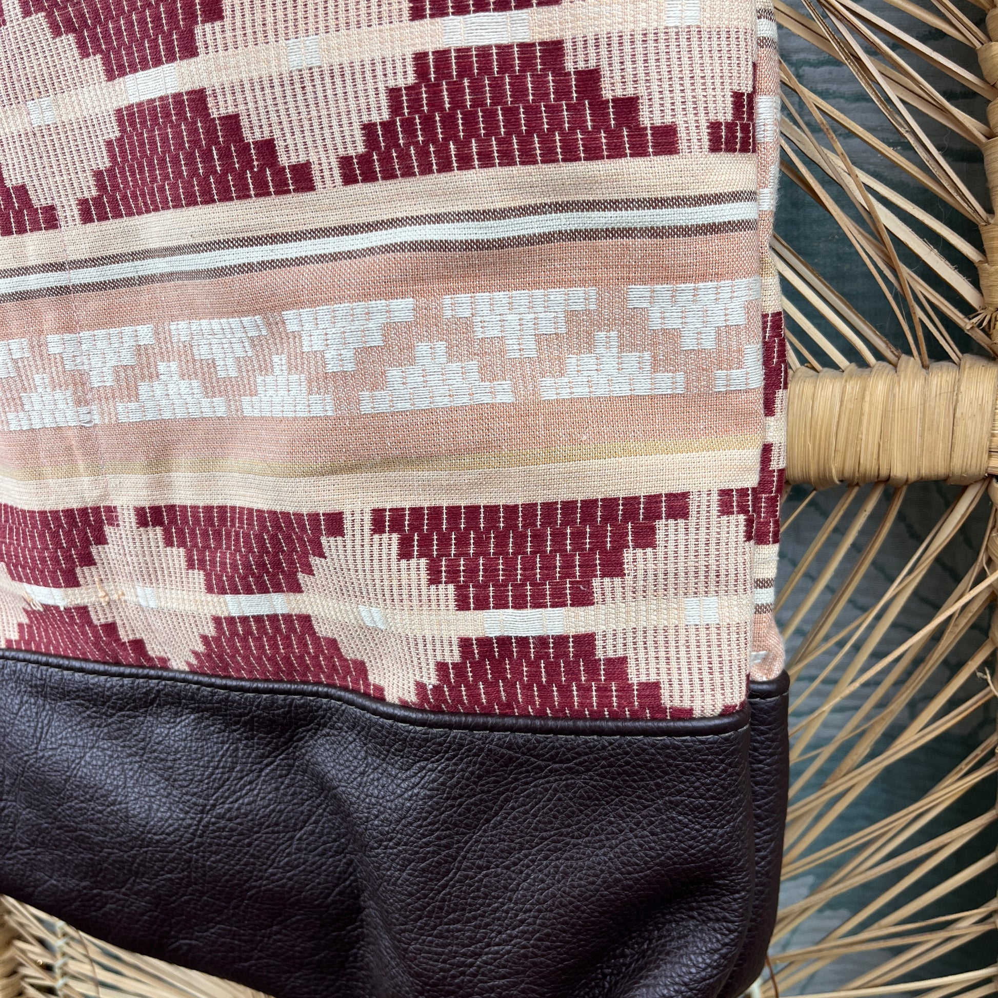 Close-up of a woven textile bag or pouch with geometric maroon, beige, and white patterns, finished with a black faux-leather bottom panel, resting on a wicker chair.