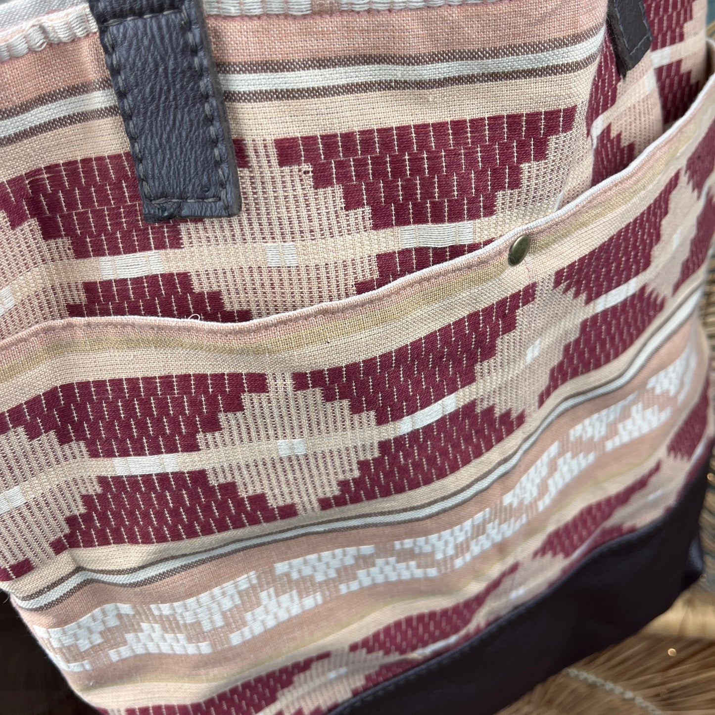 Close-up of a woven tote bag with a geometric ikat pattern in burgundy, beige, and white, featuring a front pocket and dark gray strap attachments.
